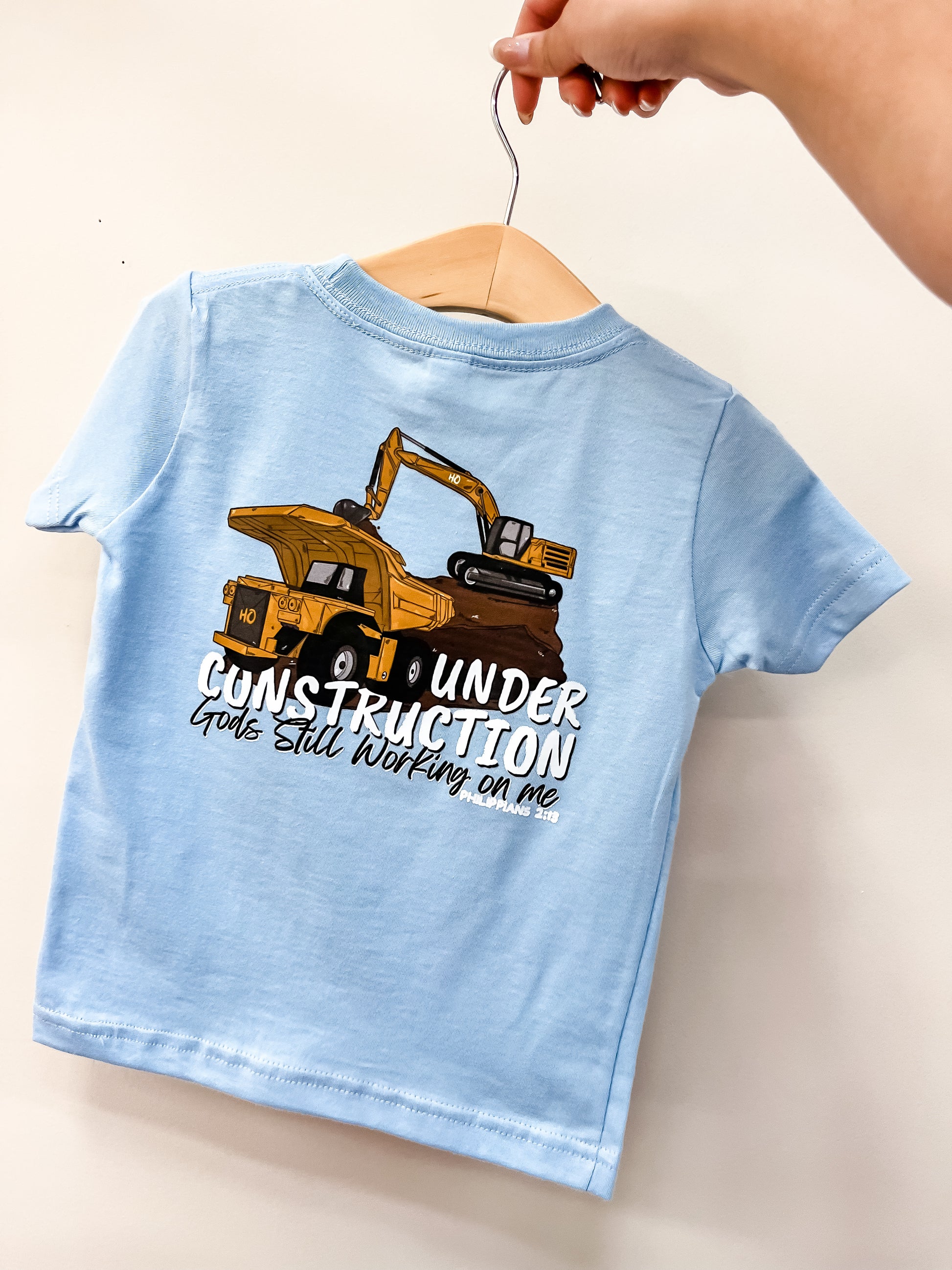 A hand holds up the hometown Outfitters Work In Progress T-Shirt for kids, light blue with a yellow excavator graphic and the words "Under Construction. God's still working on me.