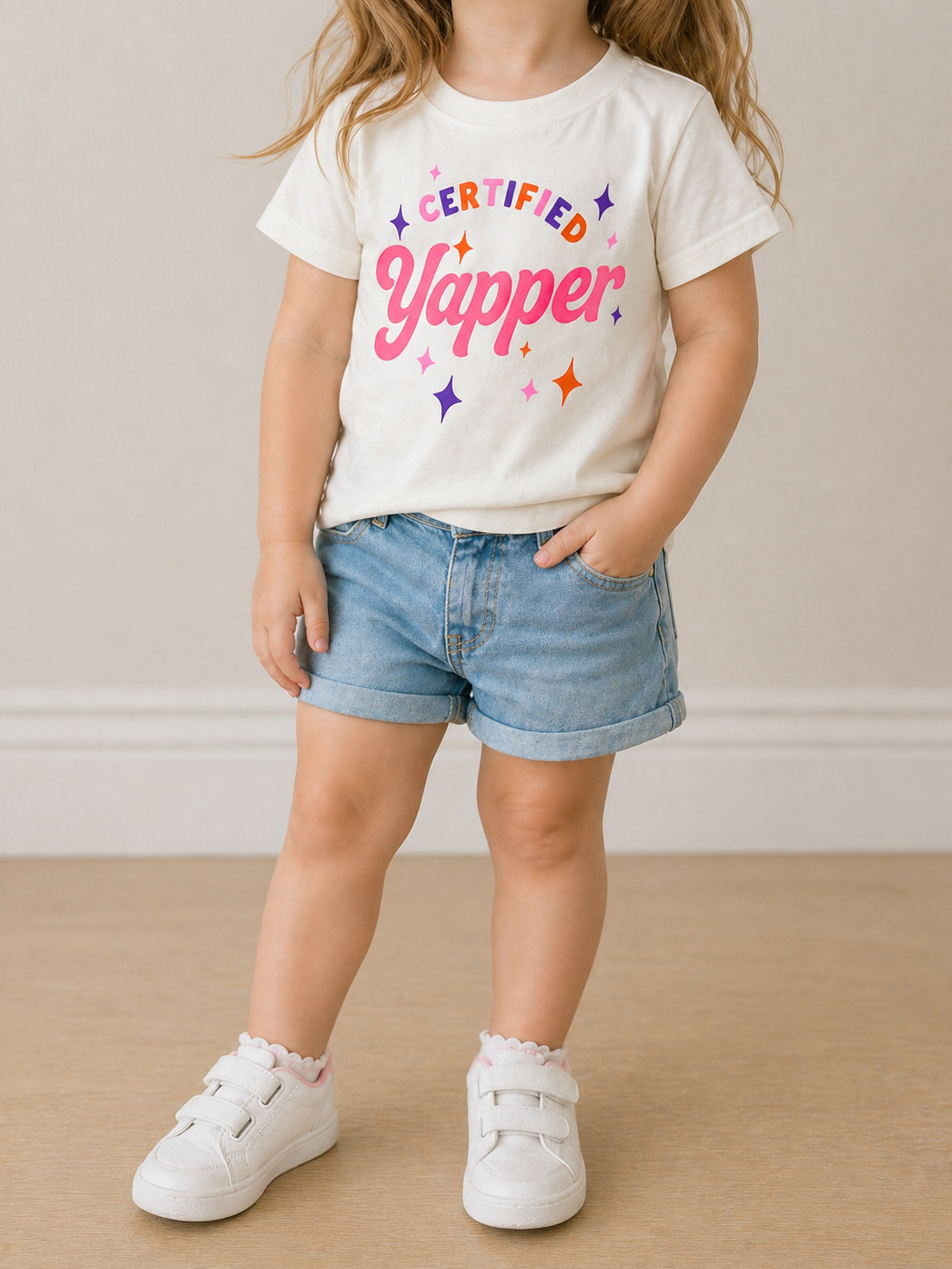 A young child poses indoors wearing the Wild Threads Co Certified Yapper T-Shirt in white, paired with denim shorts and white sneakers, with one hand in their pocket.