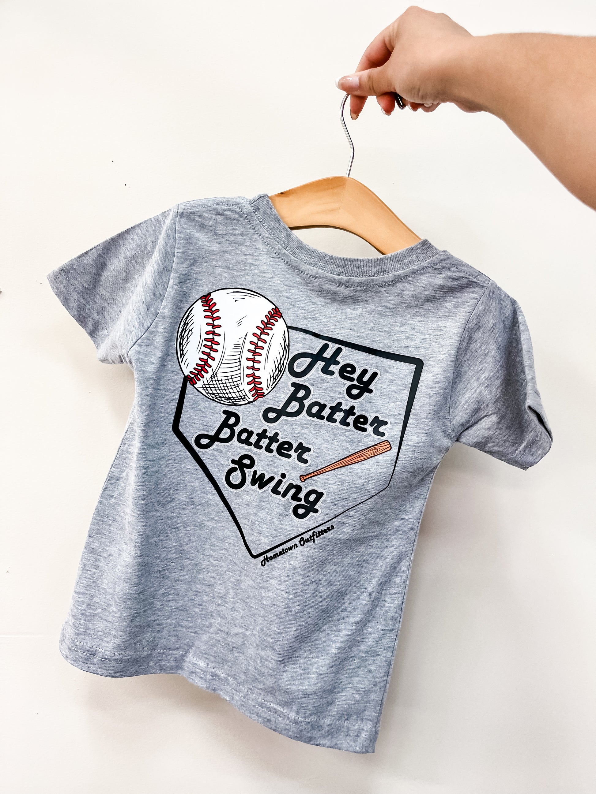 A hand displays a gray Batter Up T-Shirt by hometown Outfitters on a hanger, featuring a baseball, bat, and home plate graphic with "Hey Batter Batter Swing" printed on the back.