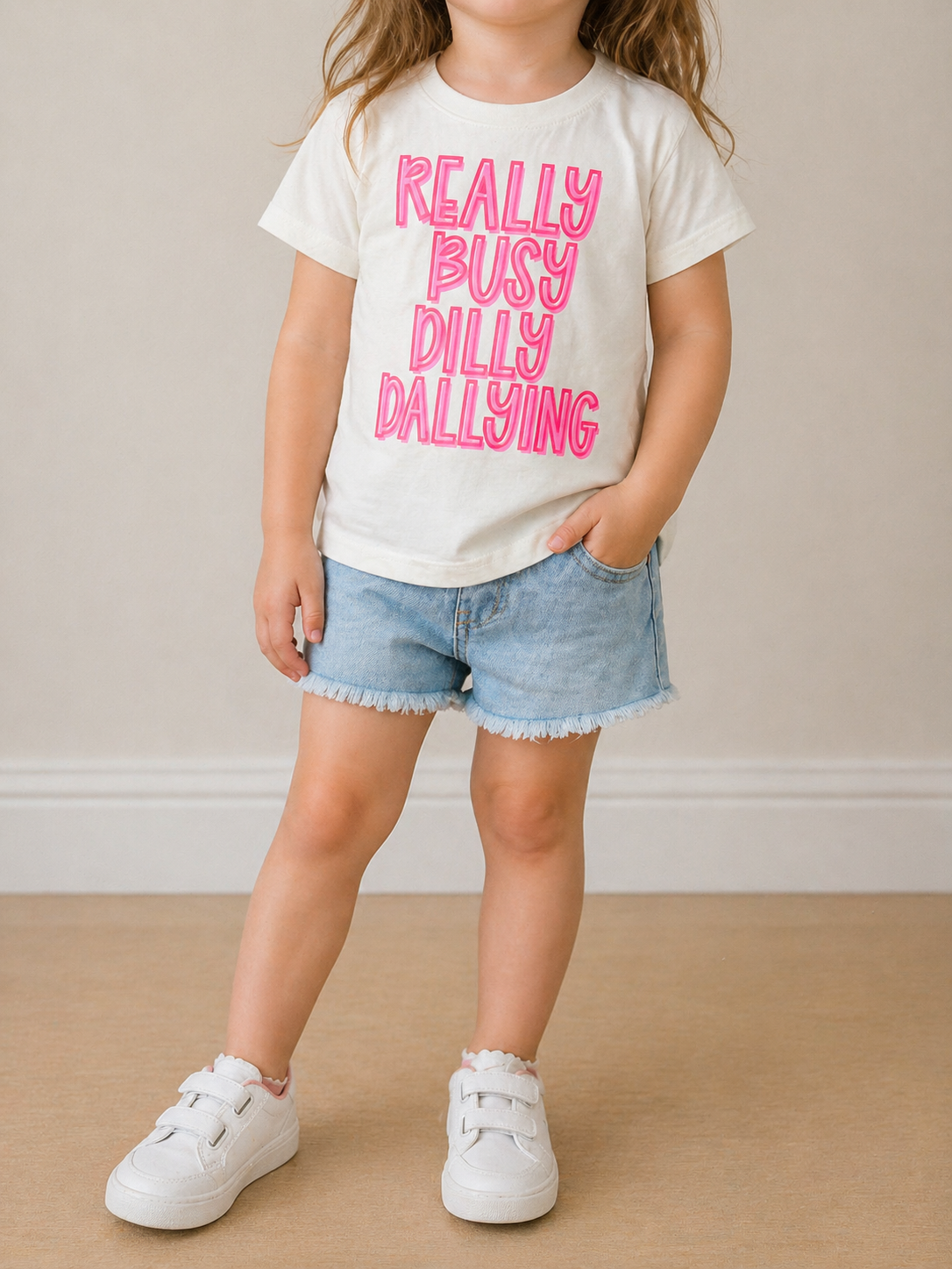 A young child wears Wild Threads Co's Dilly Dally T-Shirt with pink "Really Busy Dilly Dallying" text, paired with denim shorts and white sneakers. One hand is in their pocket against a plain, neutral background.