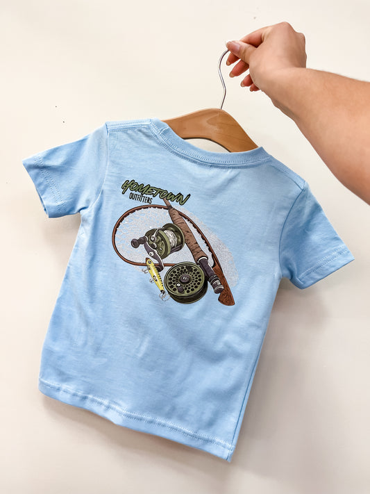 A hand holds a hanger with the Reel Cool T-Shirt by hometown Outfitters, a light blue toddler tee featuring a fishing reel and lure graphic with the brand’s name on the back.