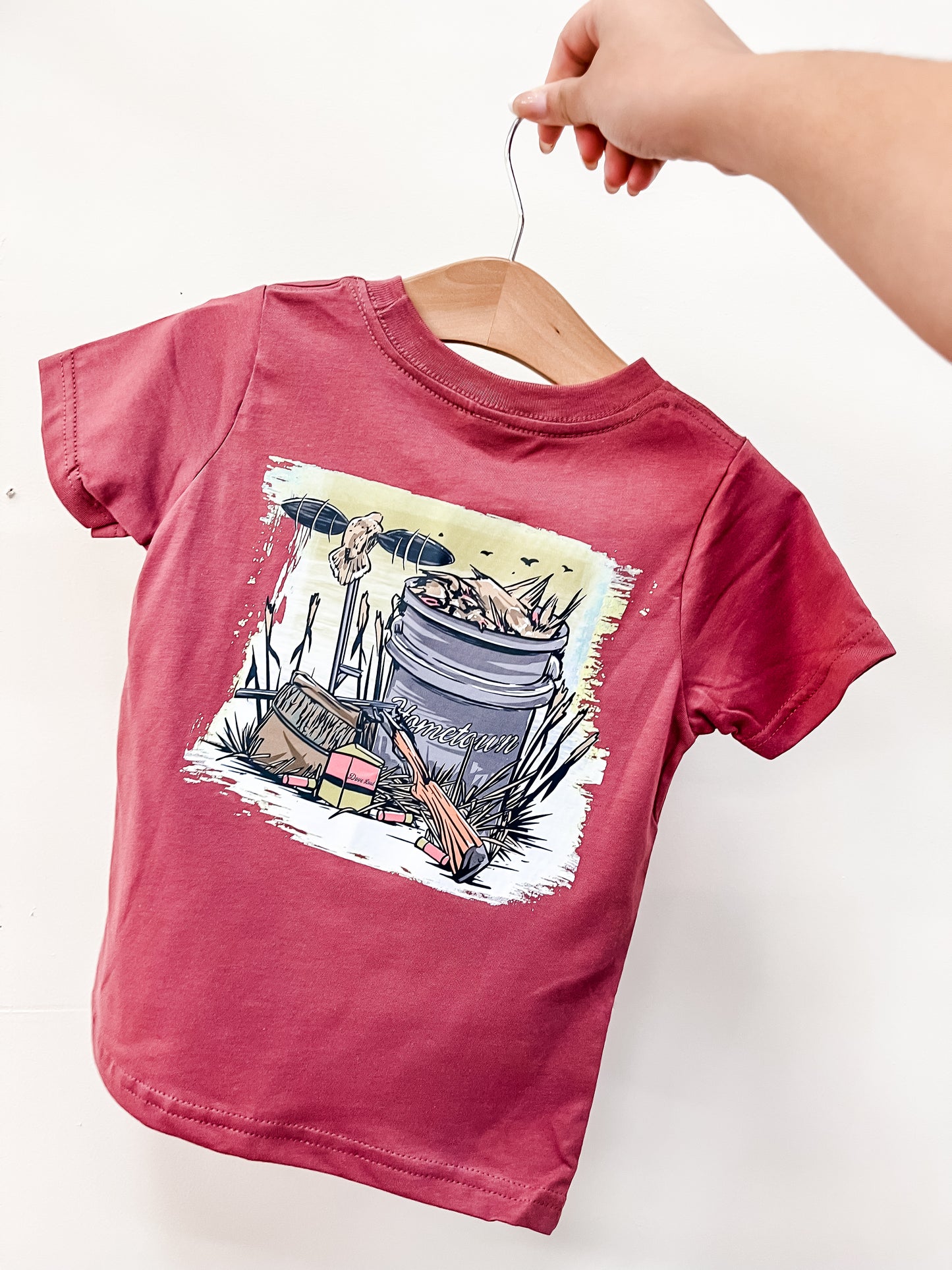 A hand holds a hanger with the Backyard Hunter T-Shirt by hometown Outfitters, a maroon children's tee featuring an outdoor-themed illustration of a bucket filled with fishing gear, reeds, tackle box, and fishing rods.