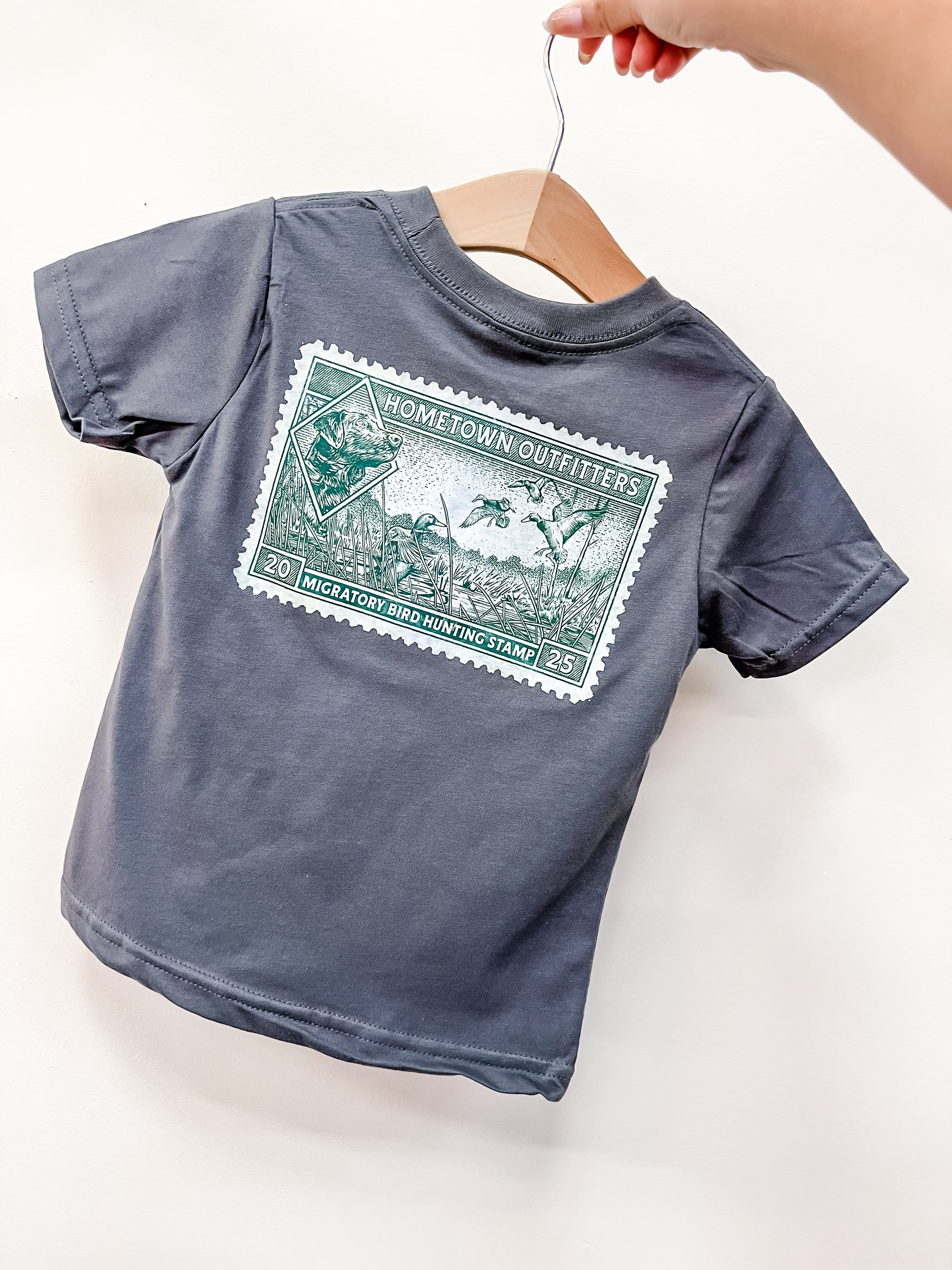 A hand holds a hanger displaying the Vintage Hunt Stamp T-Shirt by hometown Outfitters, featuring a large green “Migratory Bird Hunting Stamp 2025” graphic and hunting imagery on gray fabric.