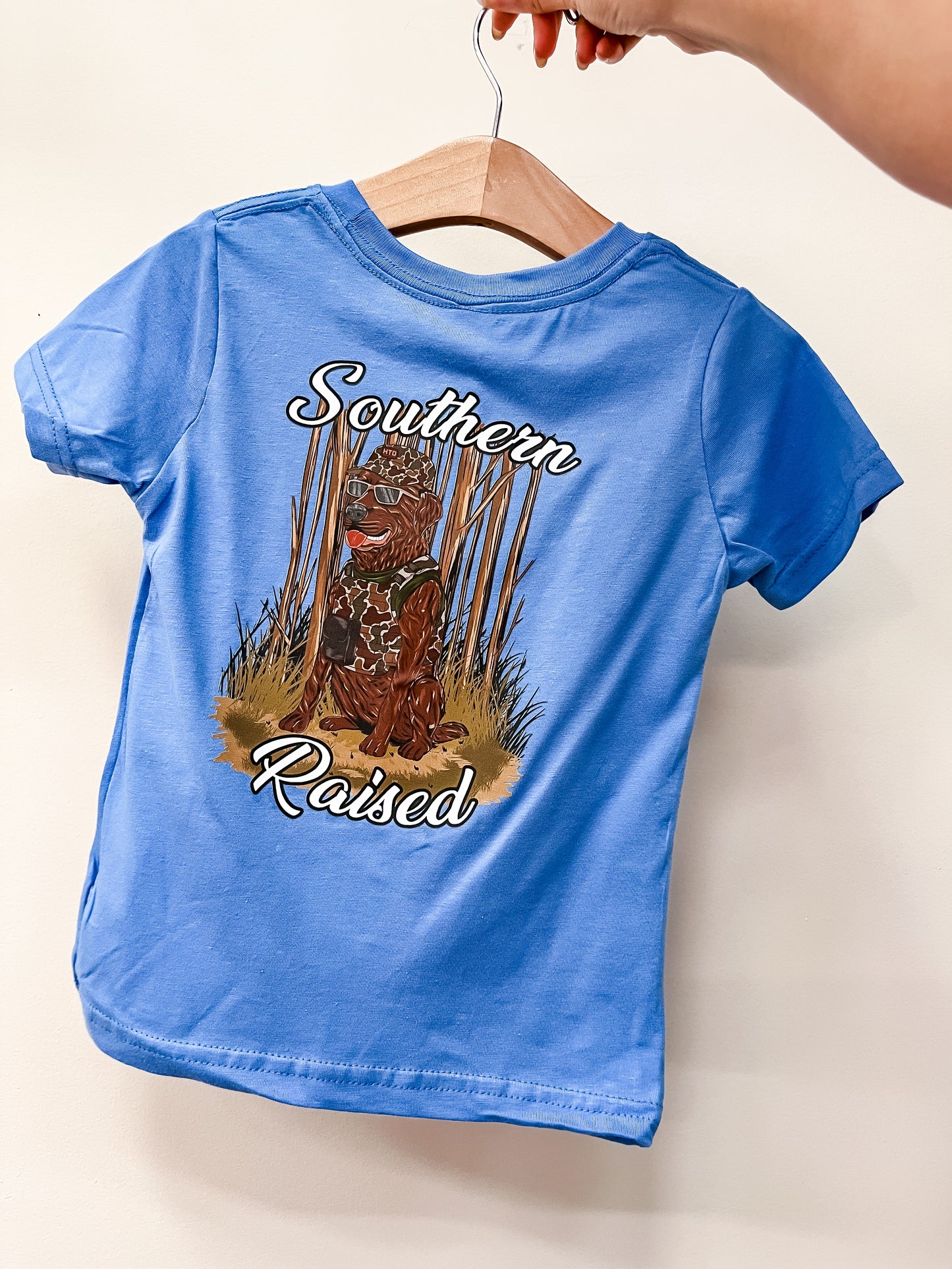 A hand holds a hanger with the Southern Raised T-Shirt by hometown Outfitters, a blue children’s shirt featuring a dog in grass and “Southern Raised” printed on the back.