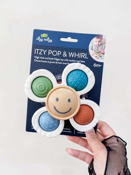 A hand holds the itzy ritzy Itzy Pop & Whirl™ - Bath + Travel Toy in its blue packaging, featuring colorful textured circles and a smiling center, designed for ages 6 months and up.
