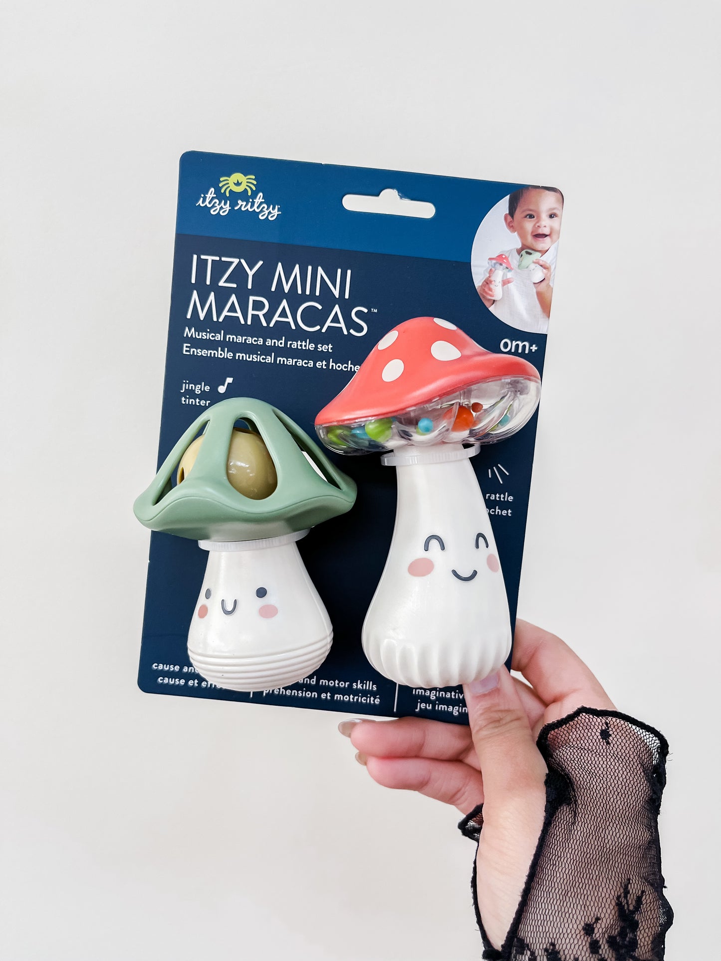 A hand holds a pack of two itzy ritzy Itzy Mini Maracas shaped like smiling mushrooms, one green and one red, against a white background. The packaging shows a baby and product details.