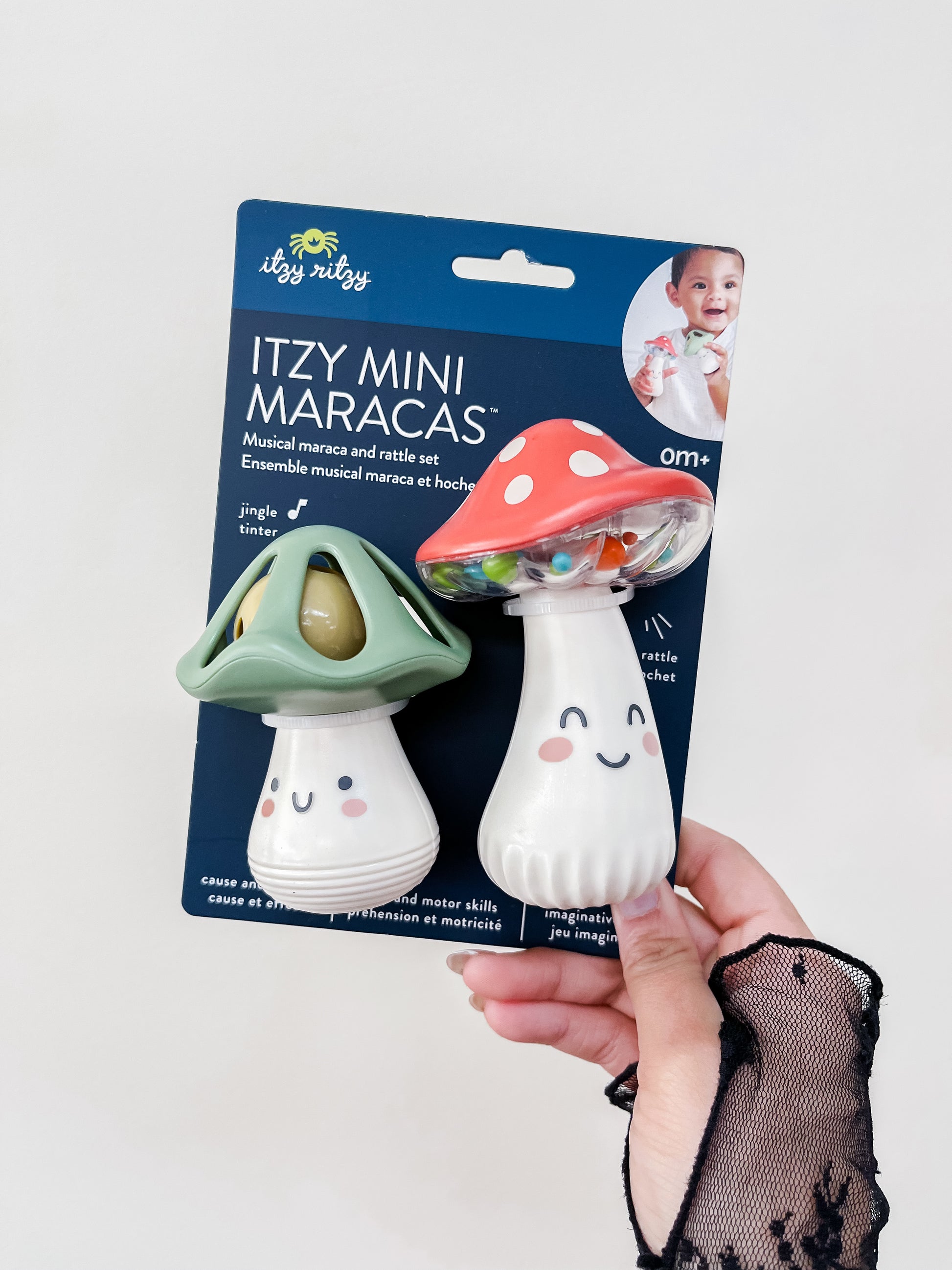 A hand holds a pack of two itzy ritzy Itzy Mini Maracas shaped like smiling mushrooms, one green and one red, against a white background. The packaging shows a baby and product details.