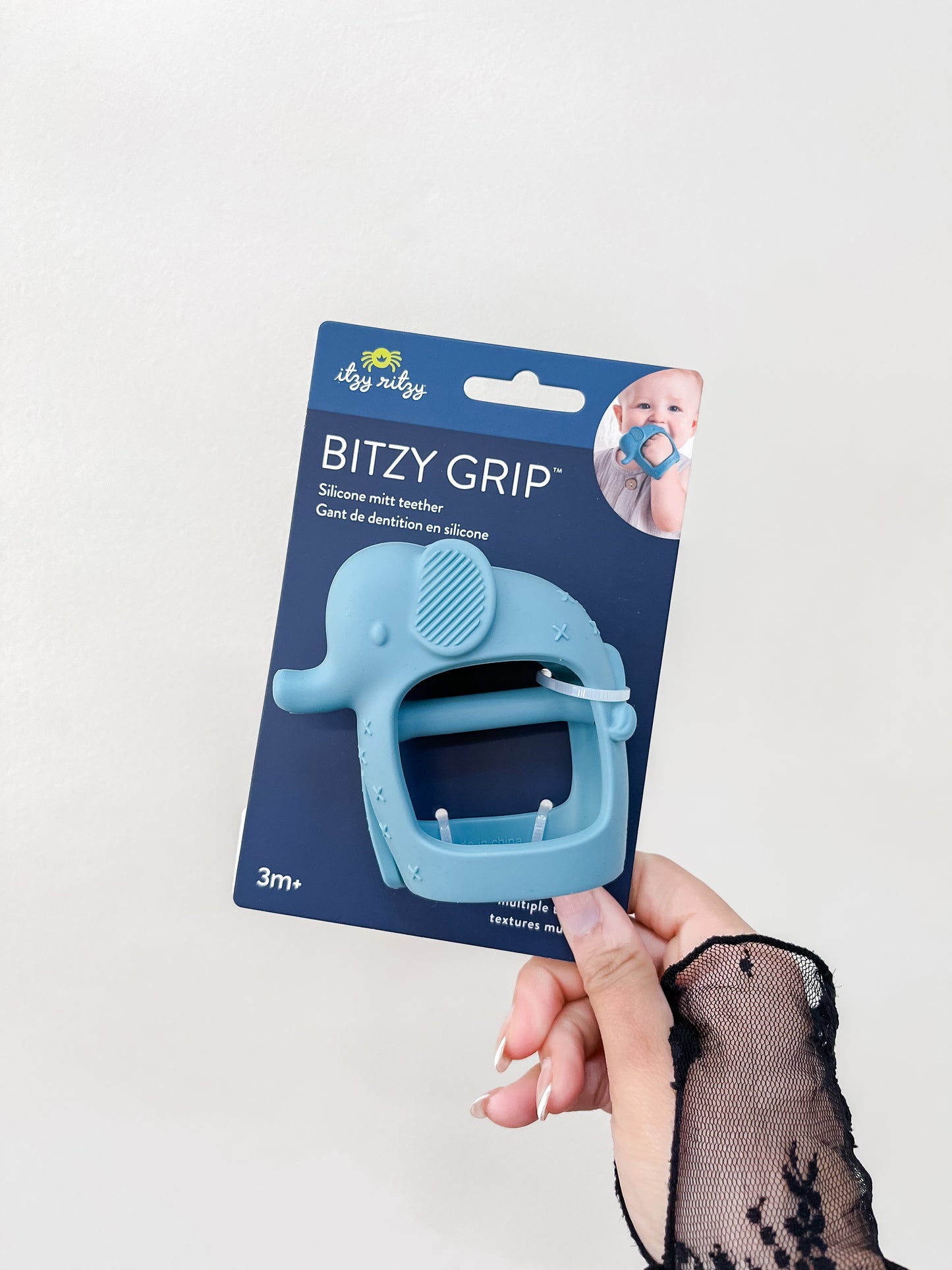 A hand with a sheer black lace sleeve holds an itzy ritzy Little Elephant Grip Toy silicone teether in packaging, designed for babies 3 months and up.