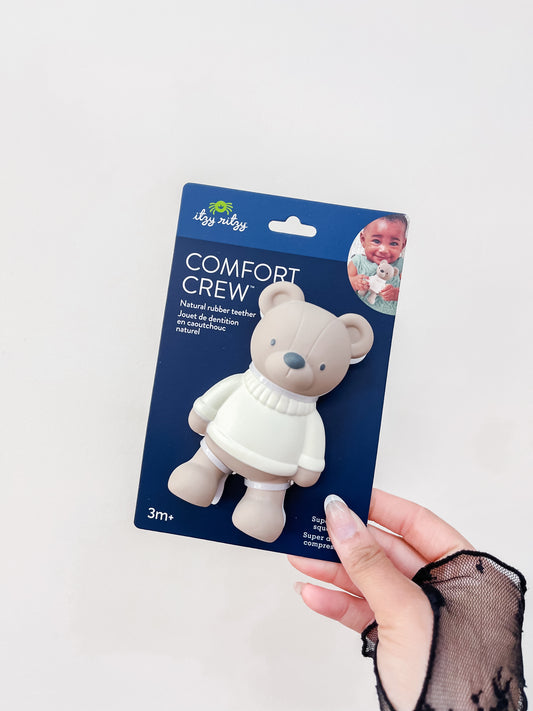 A hand with a black lace sleeve holds an itzy ritzy Cozy Bear Teether, packaged for babies aged 3 months and up. The teether is shaped like a bear in a sweater, and the packaging reads COMFORT CREW.