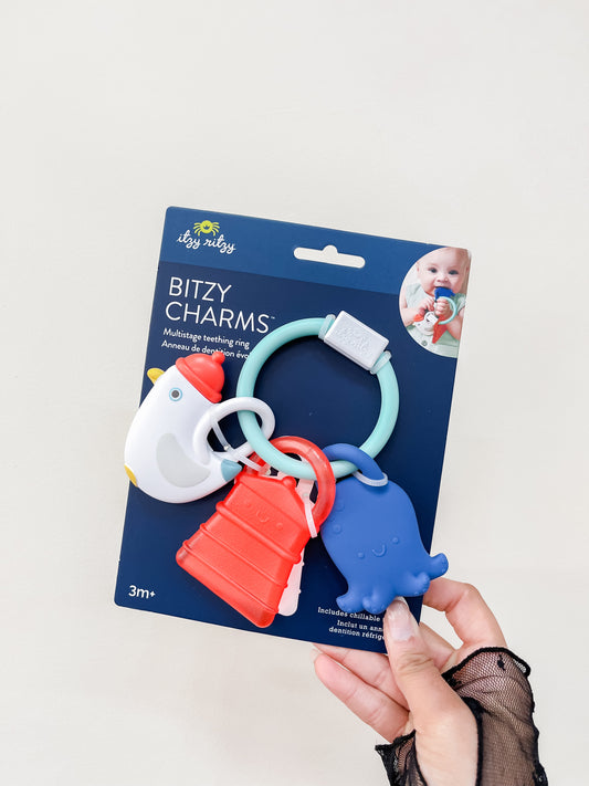 A hand holds a package of itzy ritzy Coastal Teething Charms, colorful teething rings with animal and geometric shapes for babies 3 months and up.