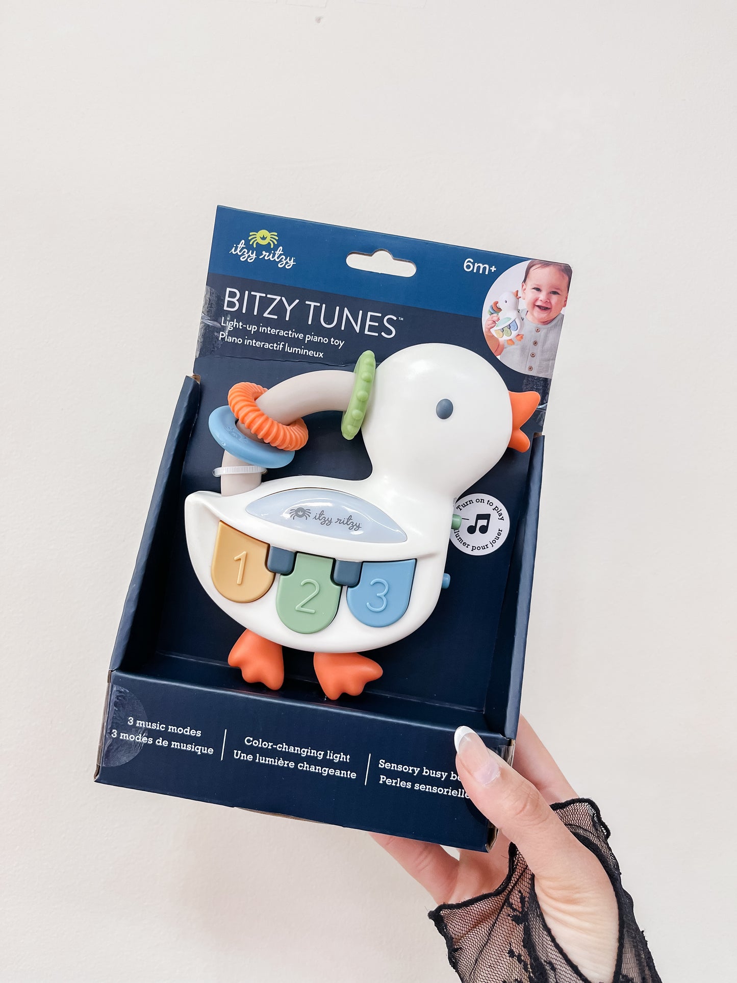 A hand holds a boxed itzy ritzy Quacky Keys Light-Up Piano, shaped like a white duck with numbered keys and colorful parts, designed for infants 6 months and up. The box displays product info and a baby’s photo.