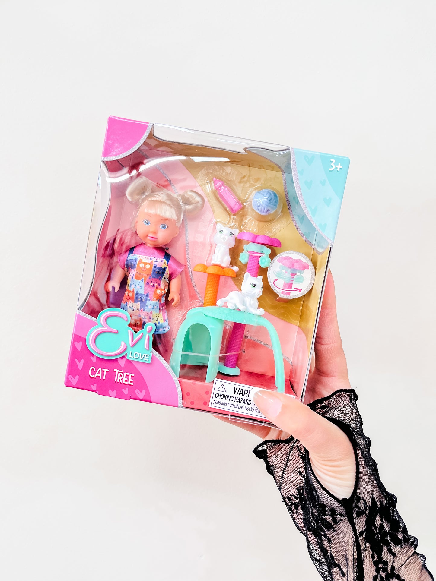 A hand in a black lace glove holds a Toy Smith Evi Love - Cat Tree Playset Doll box, featuring a doll, two white cats, a cat tree, and accessories. The packaging is decorated with pink and blue hearts.