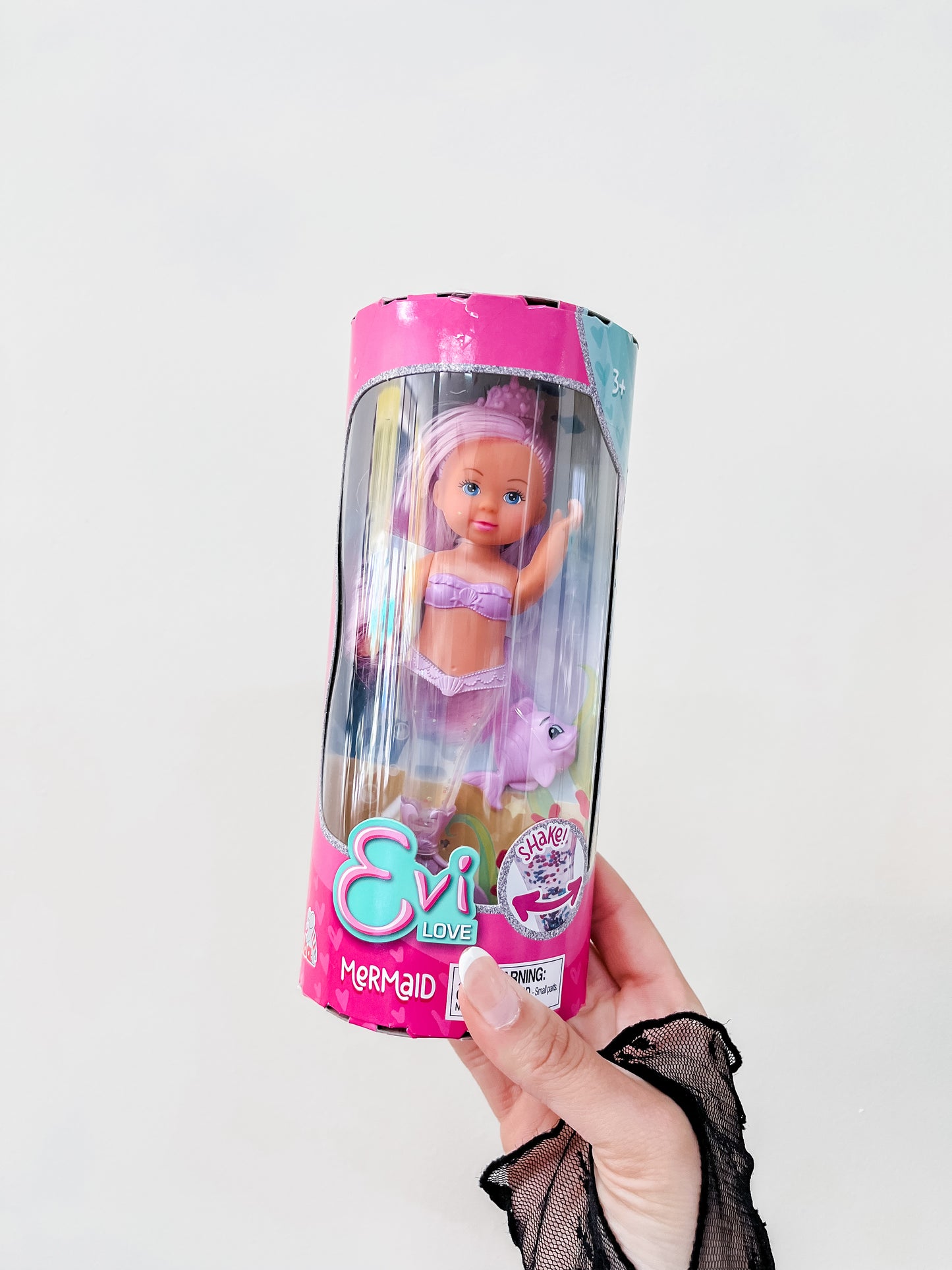 A hand in a black lace glove holds a packaged Toy Smith Evi Love Mermaid Doll with pink hair, a purple outfit, and a white unicorn figure against a plain light background.