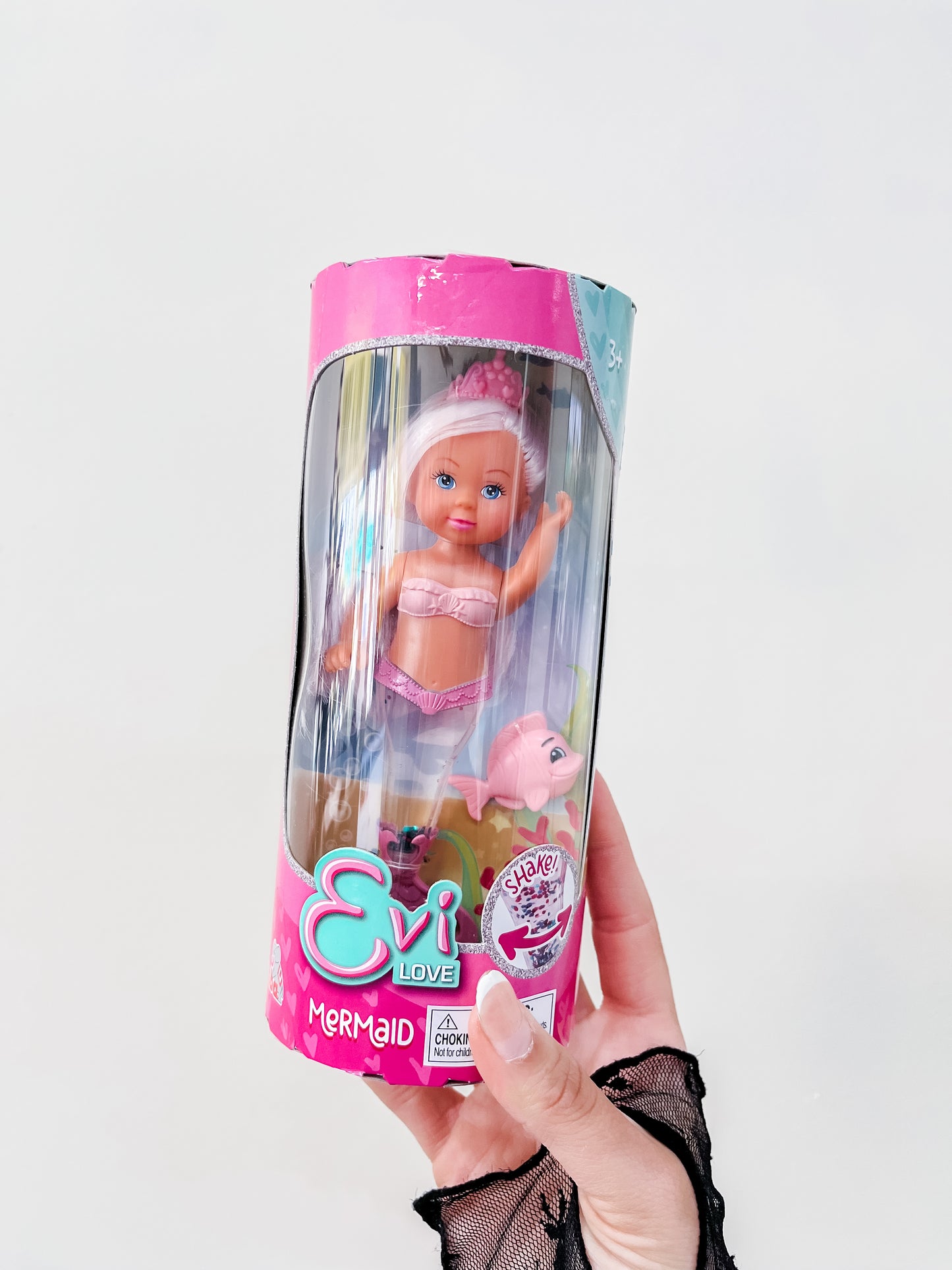 A hand holds a packaged Toy Smith Evi Love Mermaid Doll, featuring blond hair, a pink crown, pink mermaid outfit, and a small pink seahorse toy. The packaging is mainly pink and white.