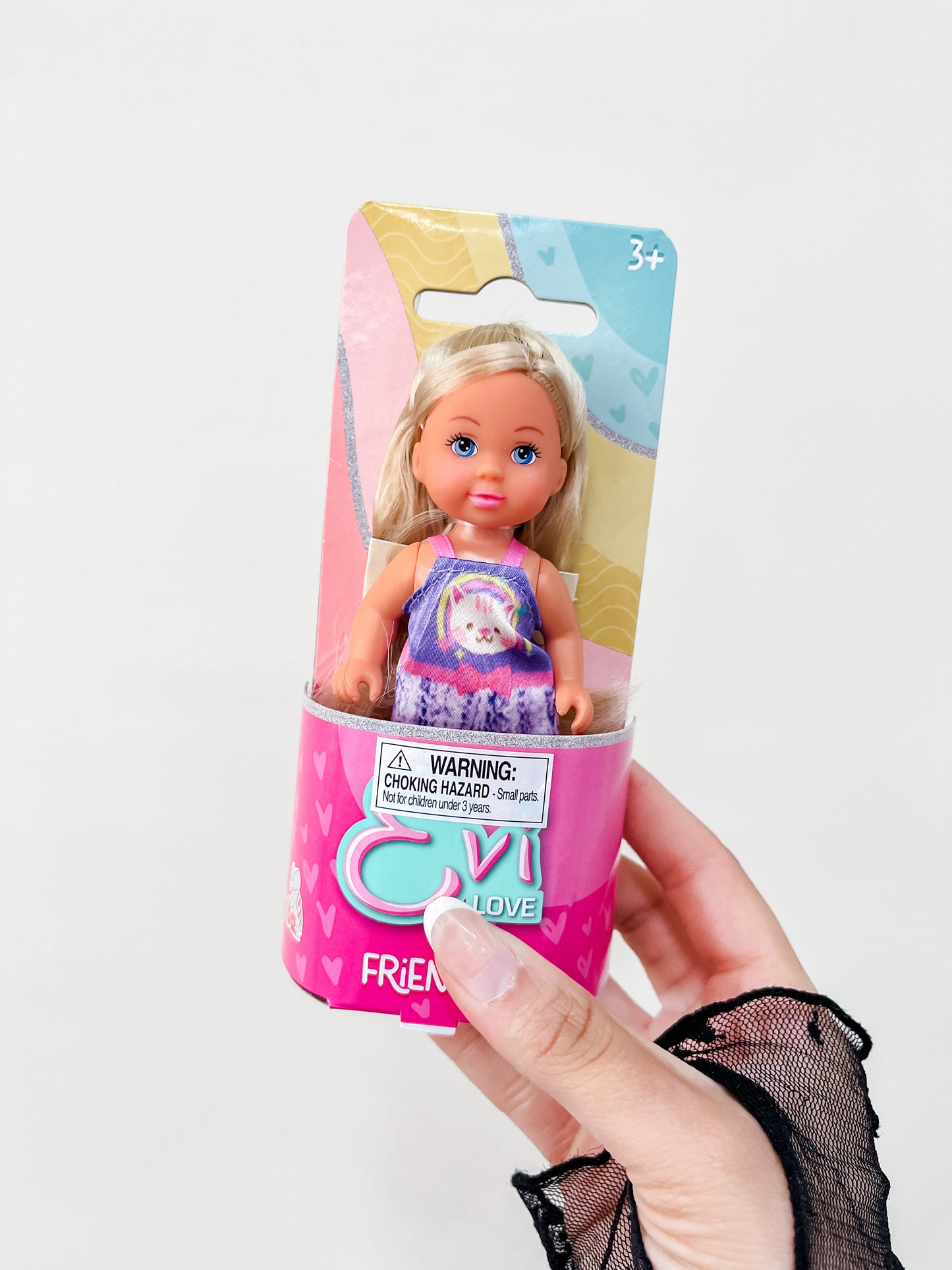 A hand holds a packaged Toy Smith Evi Love Friends Doll, featuring a blonde doll in a purple unicorn dress. The pink heart-themed packaging displays a choking hazard warning and states it's suitable for ages 3 and up.