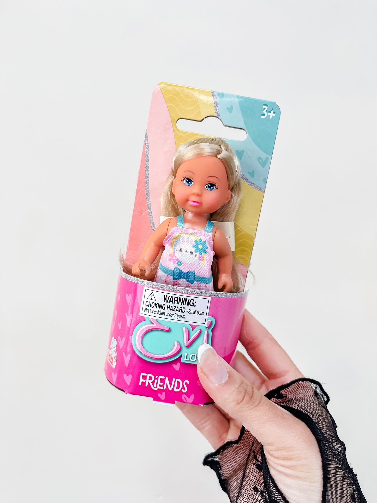 A hand with a black lace glove holds a packaged blonde Evi Love Friends Doll from Toy Smith against a plain white background. The package includes a choking hazard warning and is labeled for ages 3 and up.
