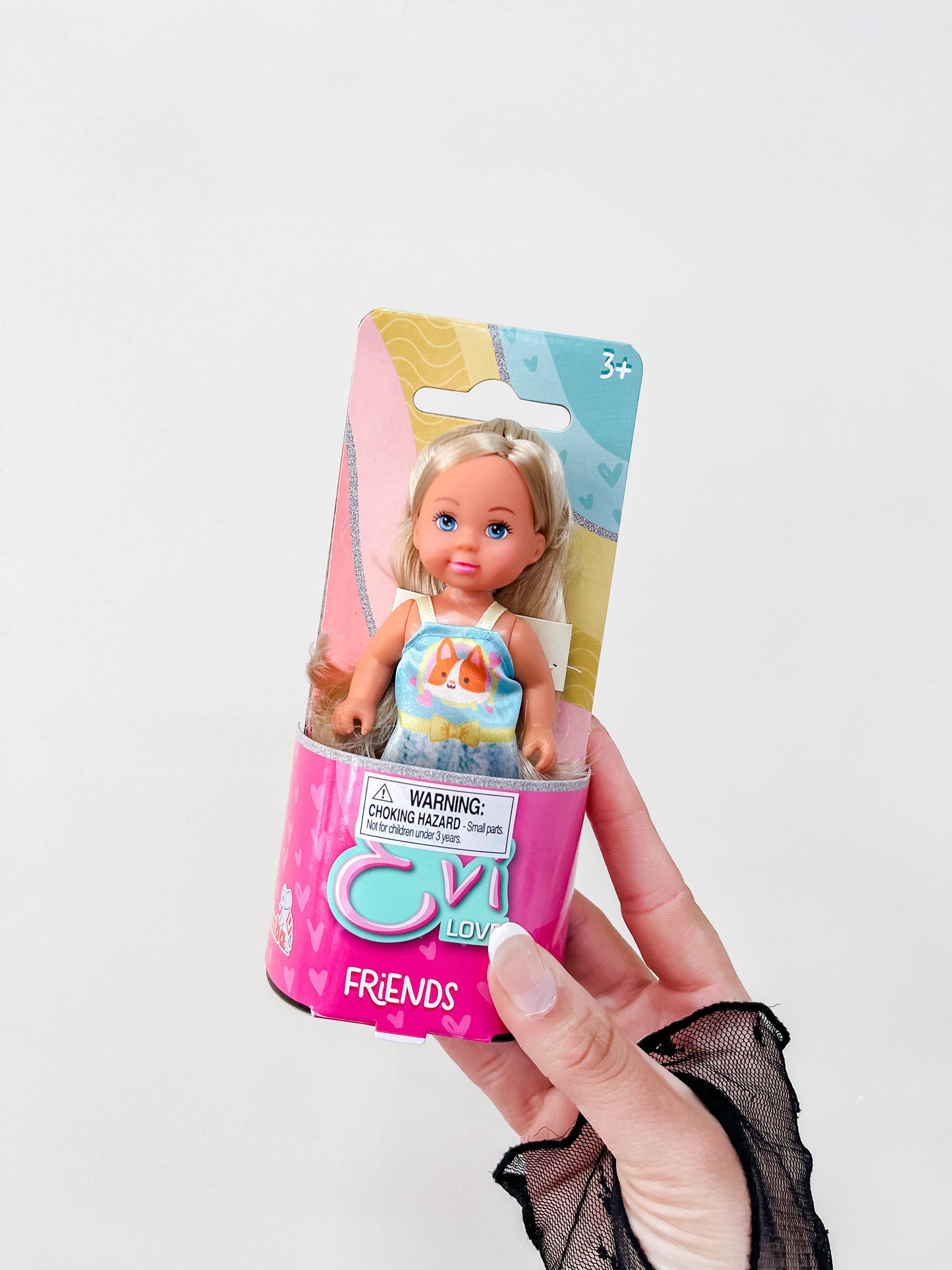A hand holds a Toy Smith Evi Love Friends Doll in bright pink packaging with playful designs and a warning label. Suitable for ages 3+, the packaging is colorful and clearly displays the product name.