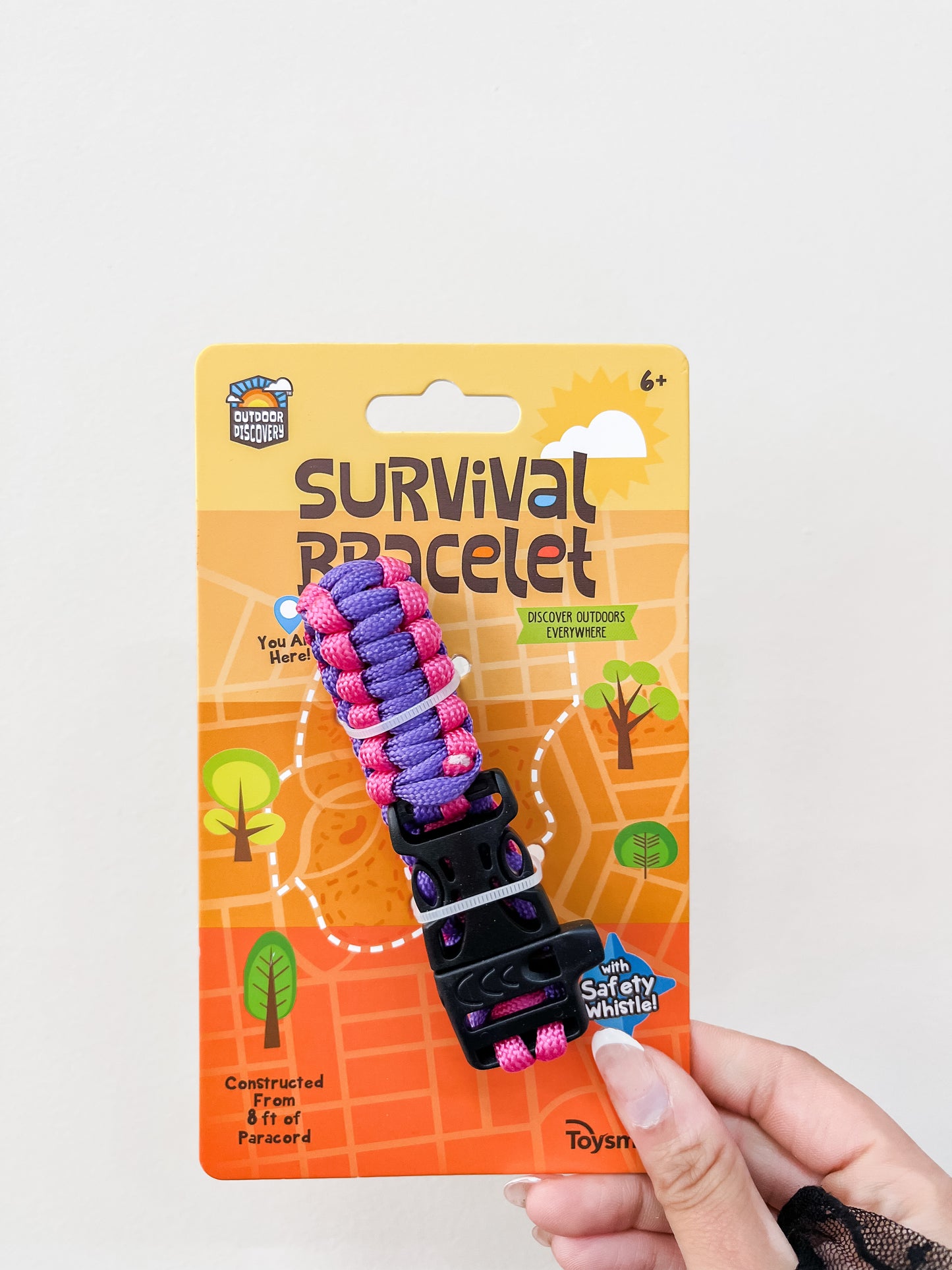 A hand holds a Toy Smith Discovery Survival Bracelet with Whistle package for ages 6+, showing outdoor icons on an orange background. The purple and pink paracord bracelet is featured, labeled "Survival Bracelet.