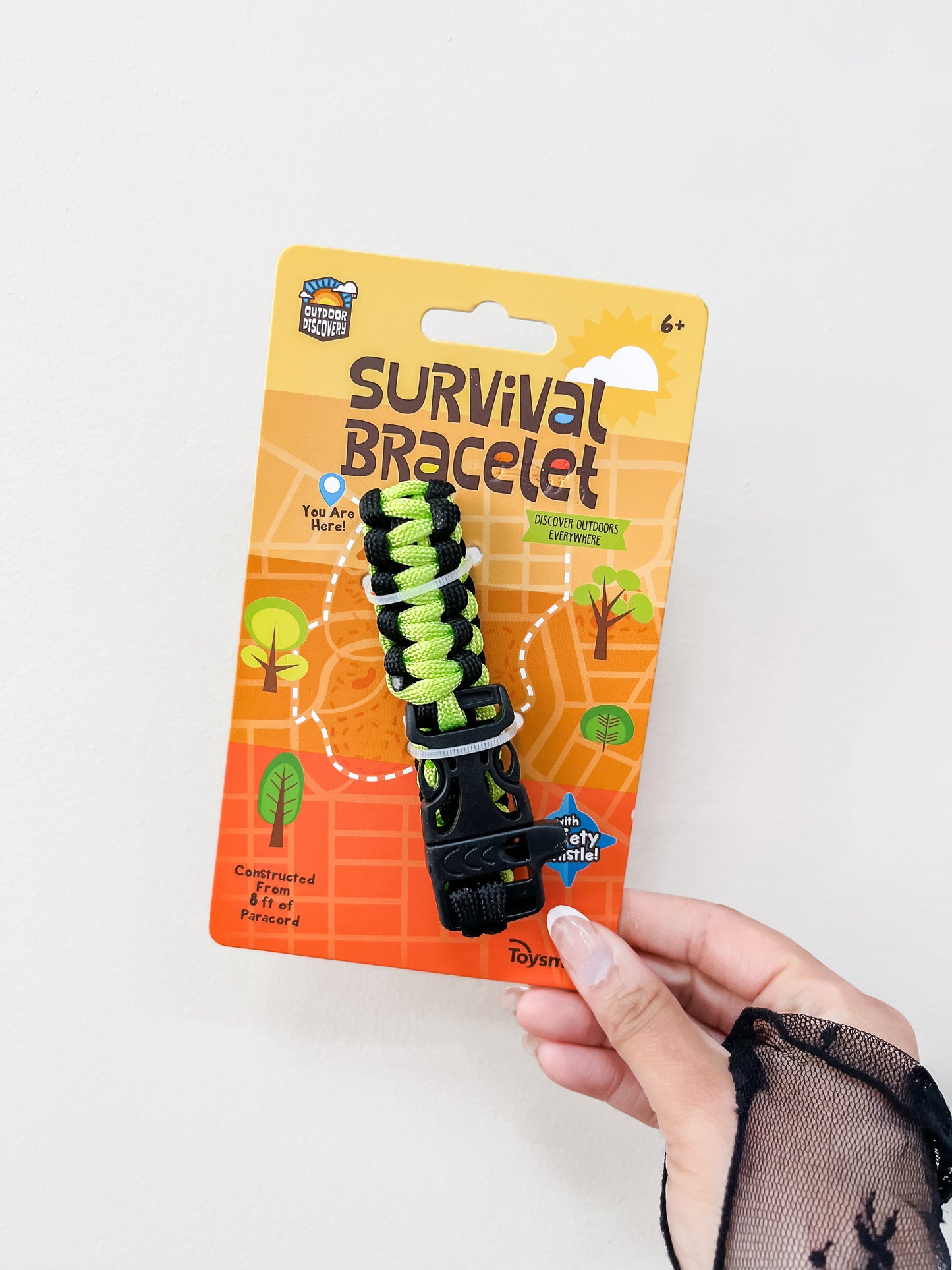A hand holds a Toy Smith Discovery Survival Bracelet with Whistle, featuring black and neon green braided cord. The orange packaging displays outdoor-themed graphics and text. Suitable for ages 6 and up.