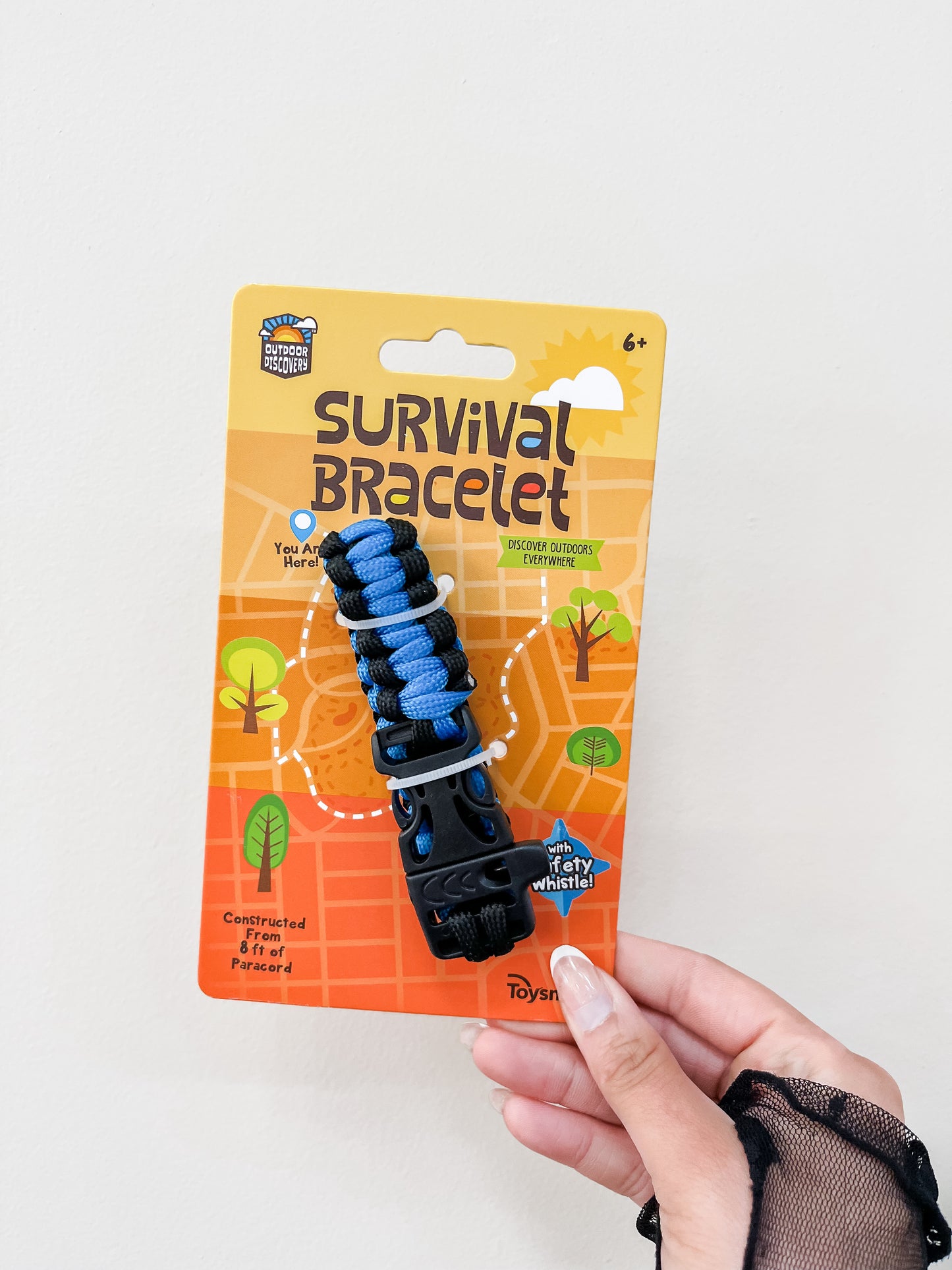 A hand holds a packaged Discovery Survival Bracelet with Whistle by Toy Smith, ages 6+, featuring blue and black colors, outdoor graphics, and package text highlighting construction features and an emergency whistle.