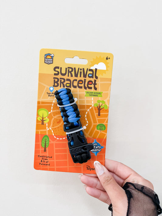 A hand holds a packaged Discovery Survival Bracelet with Whistle by Toy Smith, ages 6+, featuring blue and black colors, outdoor graphics, and package text highlighting construction features and an emergency whistle.