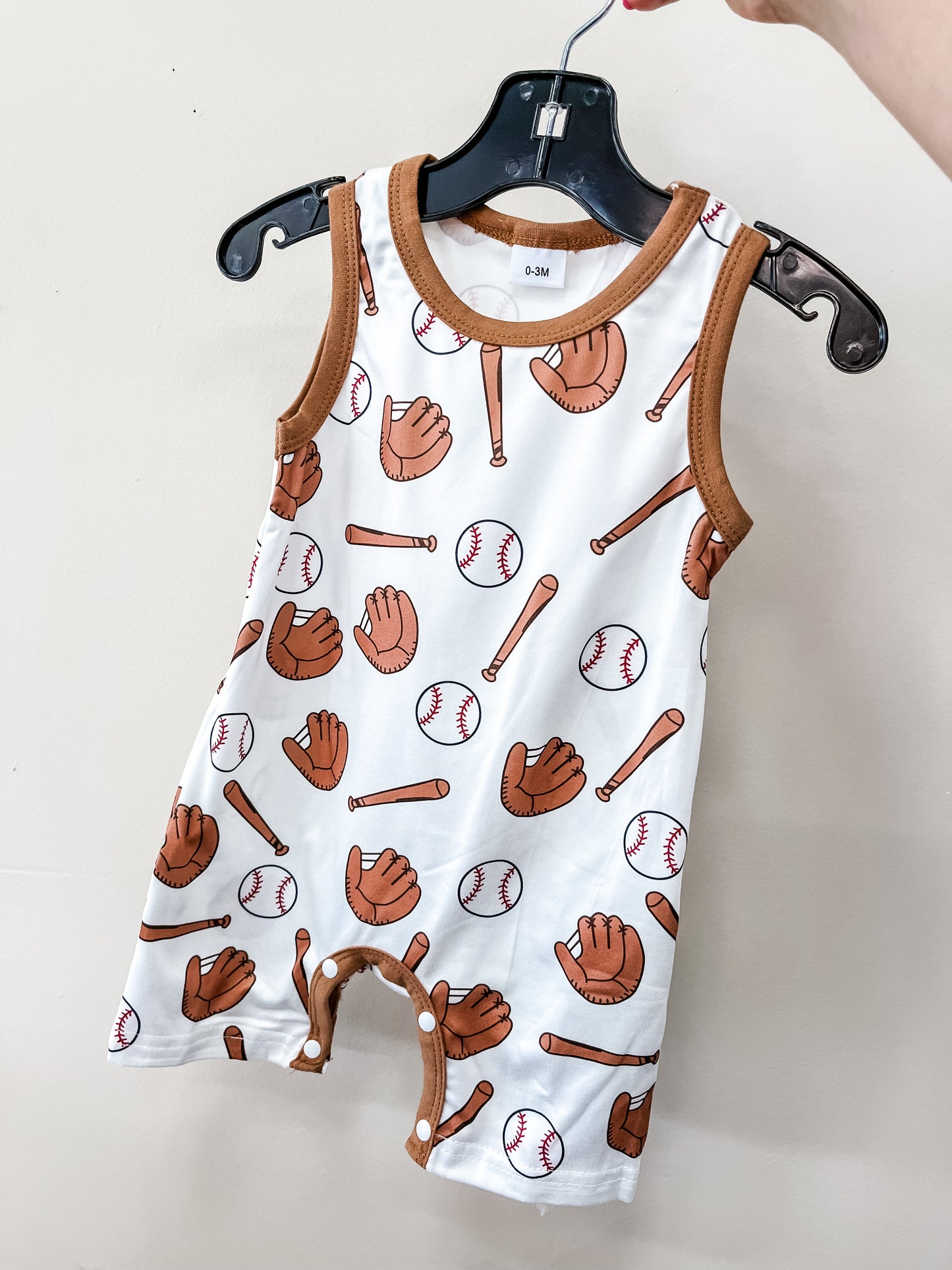 The Little Slugger Romper by Tiny Tot Bundles is a sleeveless baby outfit with brown trim and a pattern of baseballs, gloves, and bats, displayed on a black hanger against a plain light-colored wall.