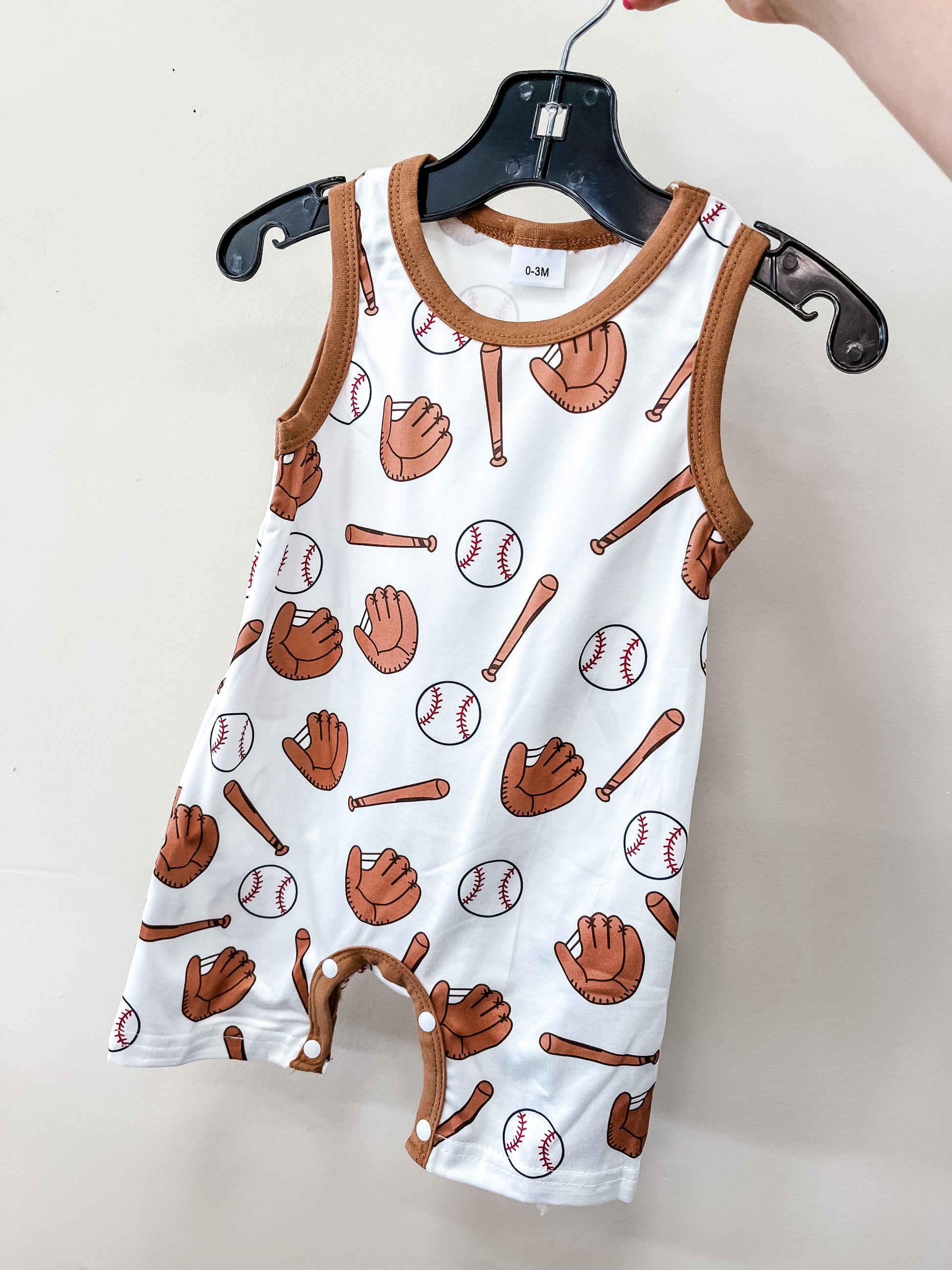 The Little Slugger Romper by Tiny Tot Bundles is a sleeveless baby outfit with brown trim and a pattern of baseballs, gloves, and bats, displayed on a black hanger against a plain light-colored wall.