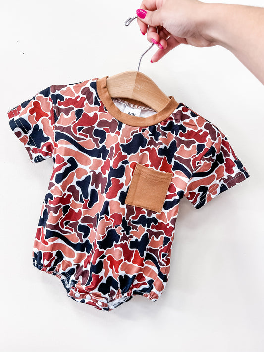 A hand with pink nails holds a Tiny Tot Bundles Rust Camo Bubble baby romper, featuring a brown, red, and black abstract camouflage pattern and a solid brown chest pocket.