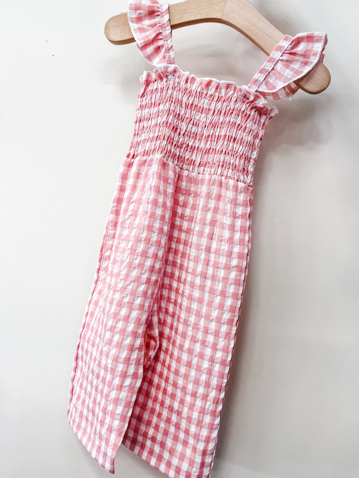 The Pink Picnic Jumpsuit by Tiny Tot Bundles, featuring pink and white gingham, ruffled shoulder straps, a smocked bodice, and a flowy skirt, hangs on a wooden hanger against a plain light wall.