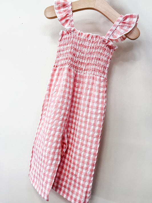 The Pink Picnic Jumpsuit by Tiny Tot Bundles, featuring pink and white gingham, ruffled shoulder straps, a smocked bodice, and a flowy skirt, hangs on a wooden hanger against a plain light wall.