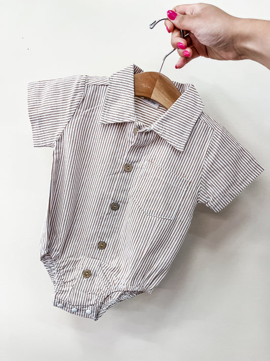 A hand with pink nail polish holds a small wooden hanger displaying the Tiny Tot Bundles Sandstone Stripe Bubble, a baby romper with short sleeves, a collar, vertical beige and white stripes, front buttons, and a chest pocket.