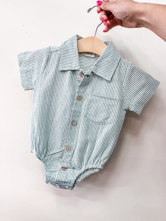 A hand holds a wooden hanger with Tiny Tot Bundles' Coastal Classic Bubble—a baby’s short-sleeved romper in light blue and white stripes, brown buttons, and a chest pocket—displayed against a plain white background.