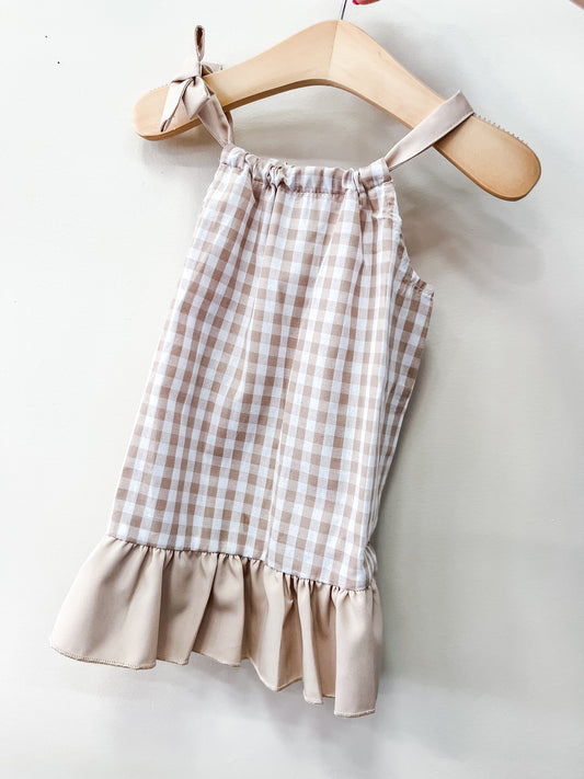 The Oatmeal Gingham Dress by Tiny Tot Bundles is a sleeveless beige and white gingham dress with shoulder ties and a ruffled hem, displayed on a wooden hanger against a white background.