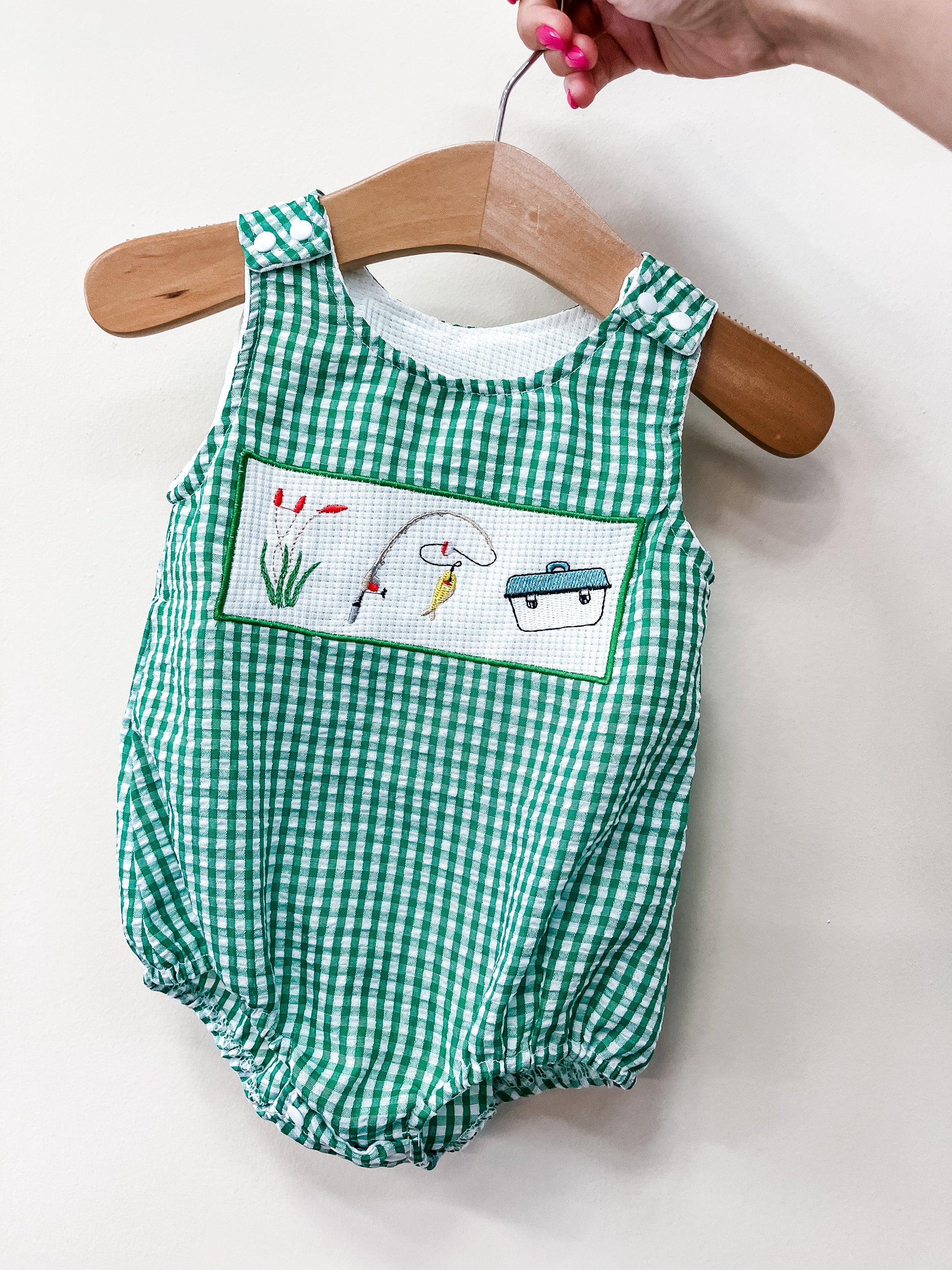 The Little Catch Bubble by Tiny Tot Bundles is a green and white gingham baby romper with fishing-themed embroidery on the front, displayed on a wooden hanger held by a hand with pink nail polish.