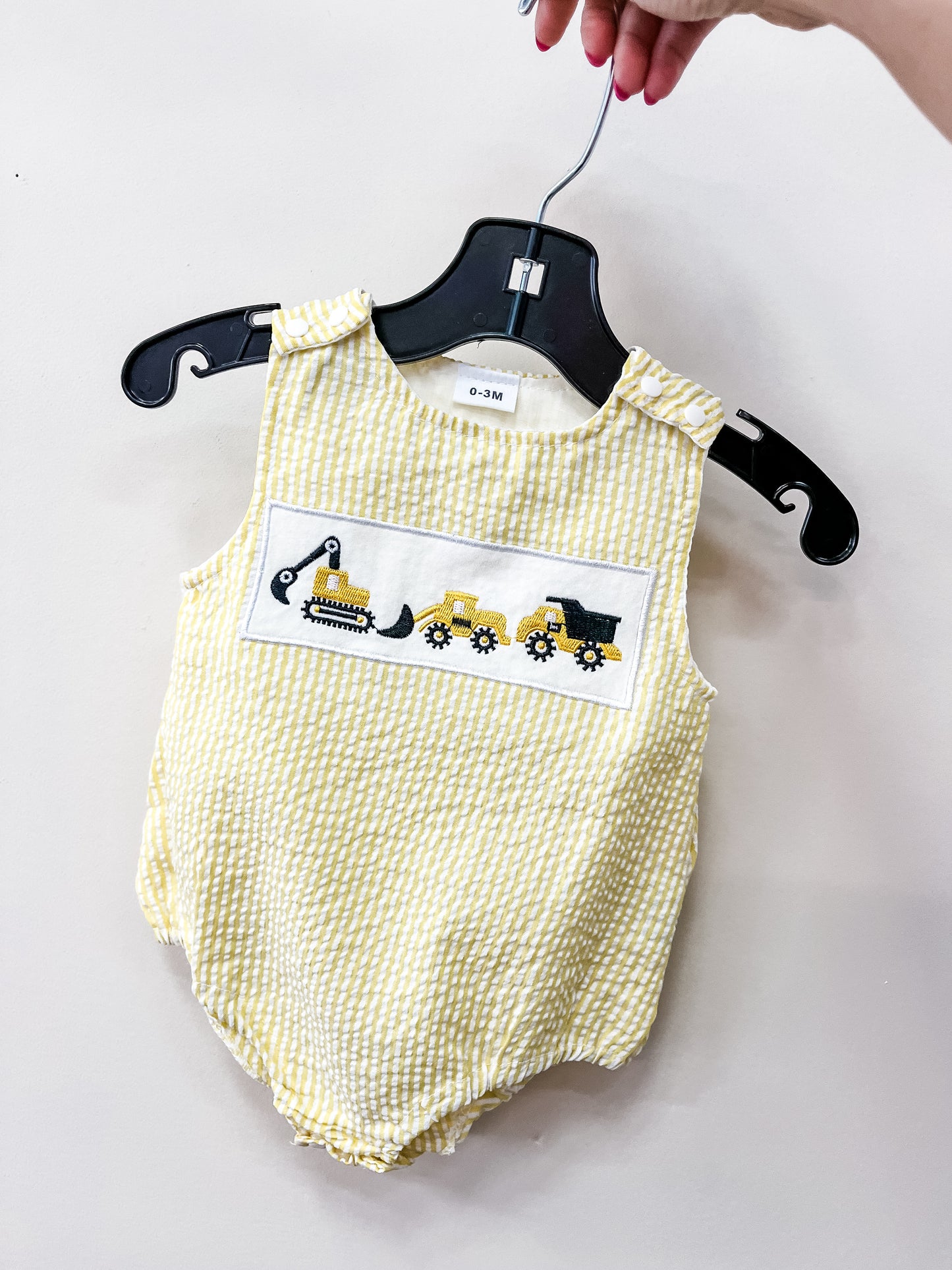 The Sunshine Builder Romper by Tiny Tot Bundles is a yellow and white checkered baby romper for ages 0-3 months, featuring embroidered construction vehicles on the chest, shown on a black hanger held against a plain background.