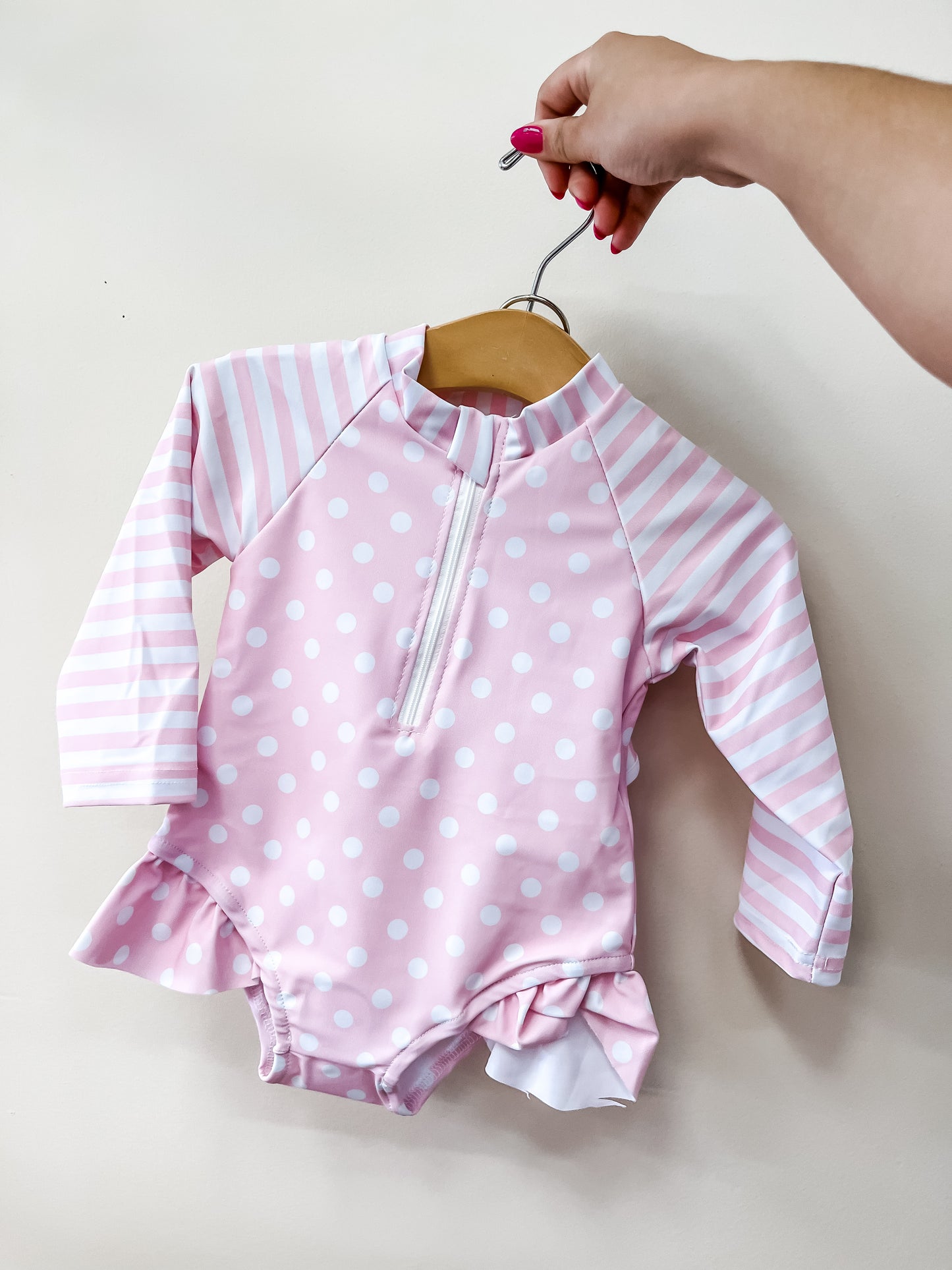 A hand holds Tiny Tot Bundles' Pink Sweet Dot Swimsuit—a baby pink suit with white polka dots, striped sleeves, front zipper, and ruffled legs—against a plain light background.