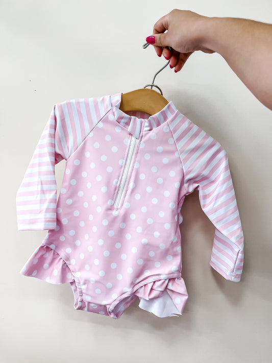 A hand holds Tiny Tot Bundles' Pink Sweet Dot Swimsuit—a baby pink suit with white polka dots, striped sleeves, front zipper, and ruffled legs—against a plain light background.