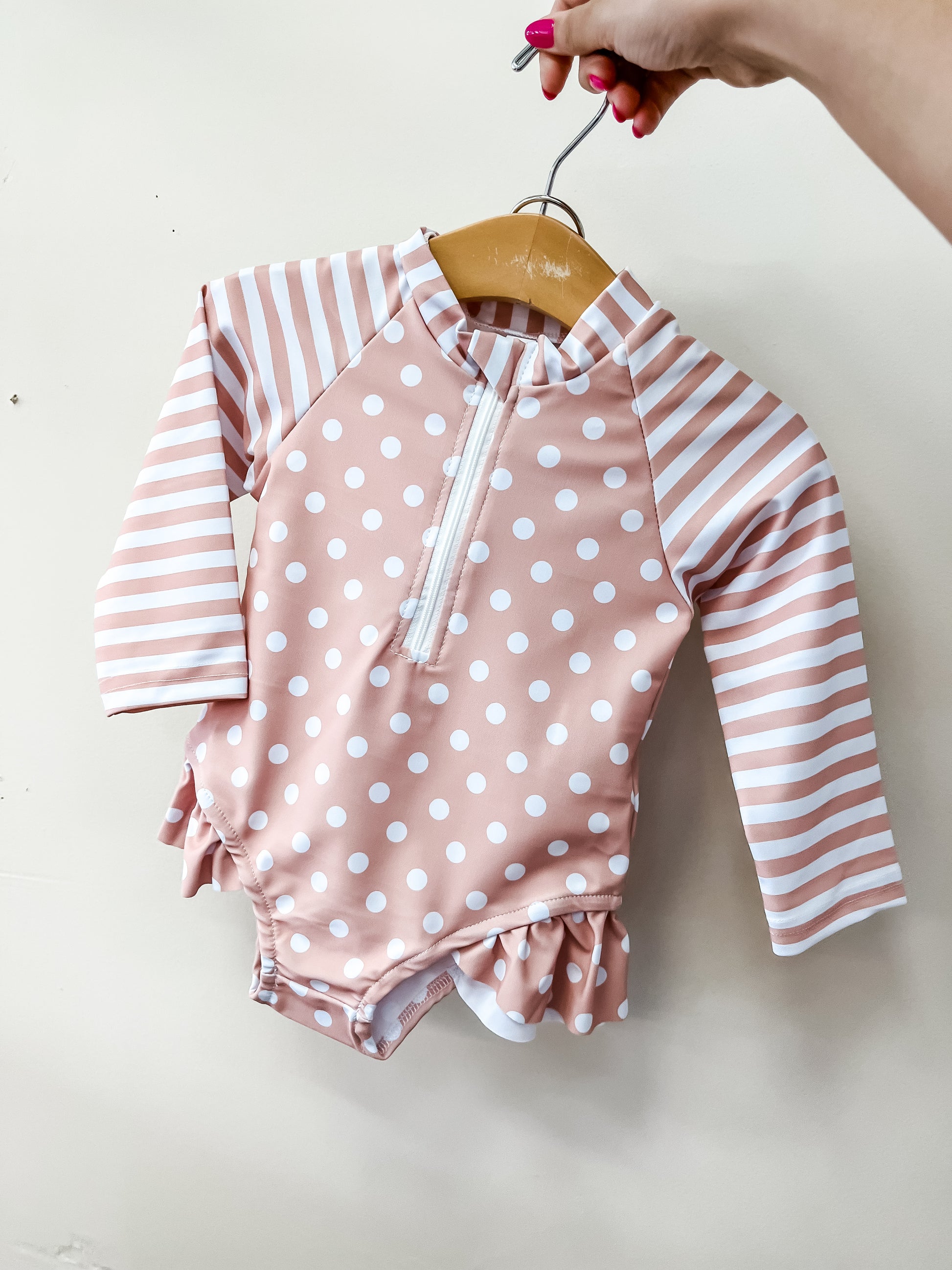 A hand holds the Tiny Tot Bundles Beige Sweet Dot Swimsuit, featuring white dots on the body and horizontal stripes on the long sleeves, against a plain light background.