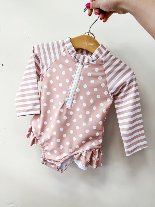 A hand holds the Tiny Tot Bundles Beige Sweet Dot Swimsuit, featuring white dots on the body and horizontal stripes on the long sleeves, against a plain light background.