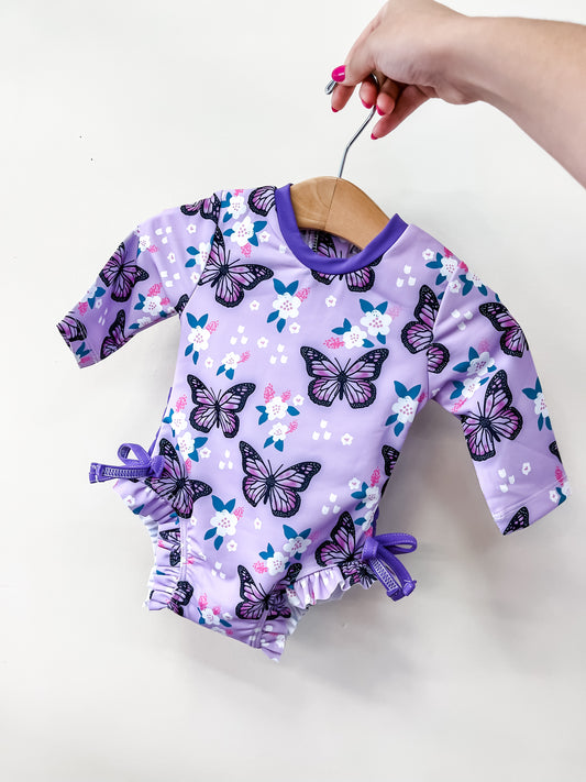 A hand holds the Tiny Tot Bundles Butterfly Kisses Swimsuit—a purple baby romper with black and pink butterflies, white and blue flowers, ruffled leg openings, and purple bows—against a plain light background.