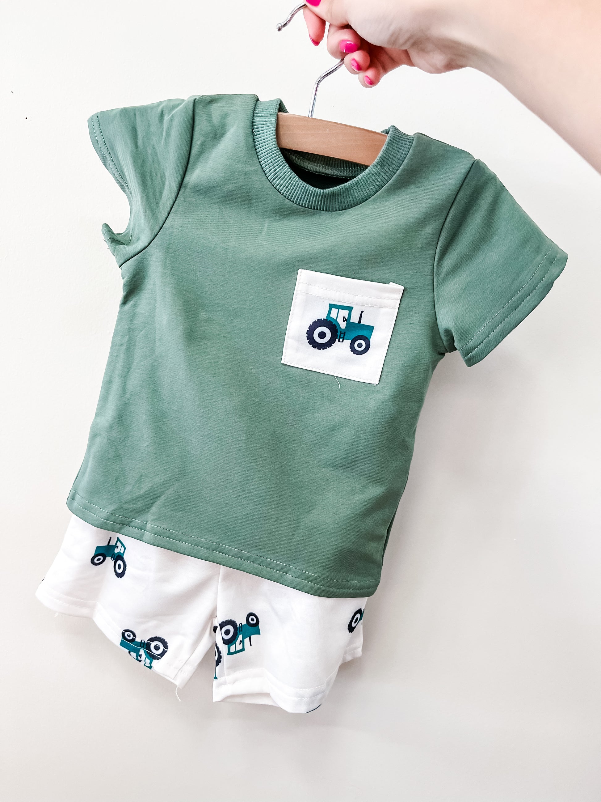 The Little Farmer Set by Tiny Tot Bundles features a green toddler shirt with a blue tractor pocket graphic and matching white shorts with blue tractor prints, displayed on a wooden hanger held by a hand with pink painted nails.