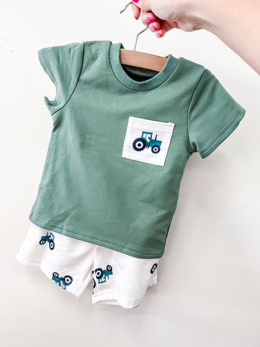 The Little Farmer Set by Tiny Tot Bundles features a green toddler shirt with a blue tractor pocket graphic and matching white shorts with blue tractor prints, displayed on a wooden hanger held by a hand with pink painted nails.