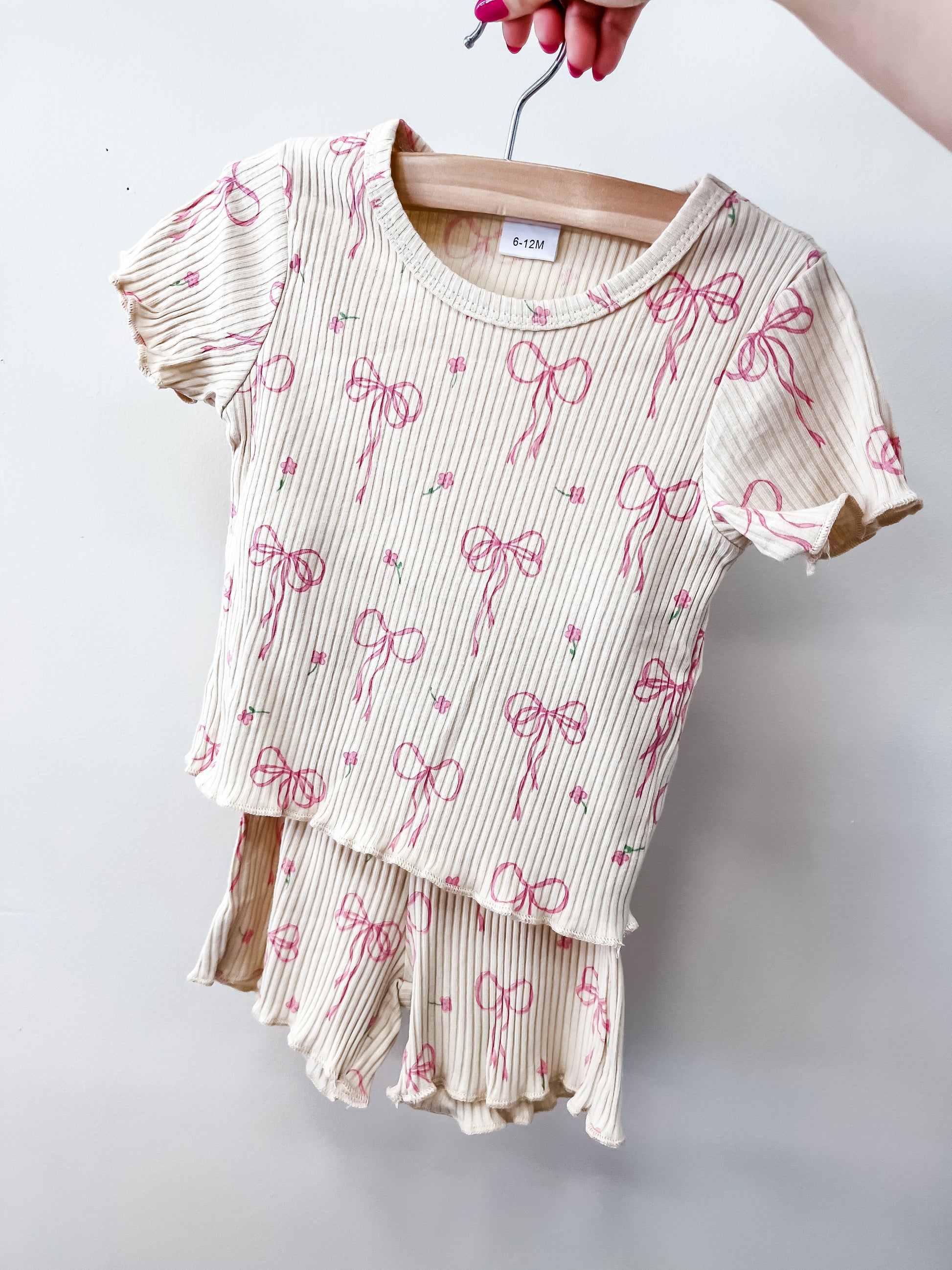 A hand displays the Sugar Bow Set by Tiny Tot Bundles—a cream-colored, ribbed two-piece kids' outfit with pink bows and green dots. The set includes a short-sleeve top and matching ruffled shorts.