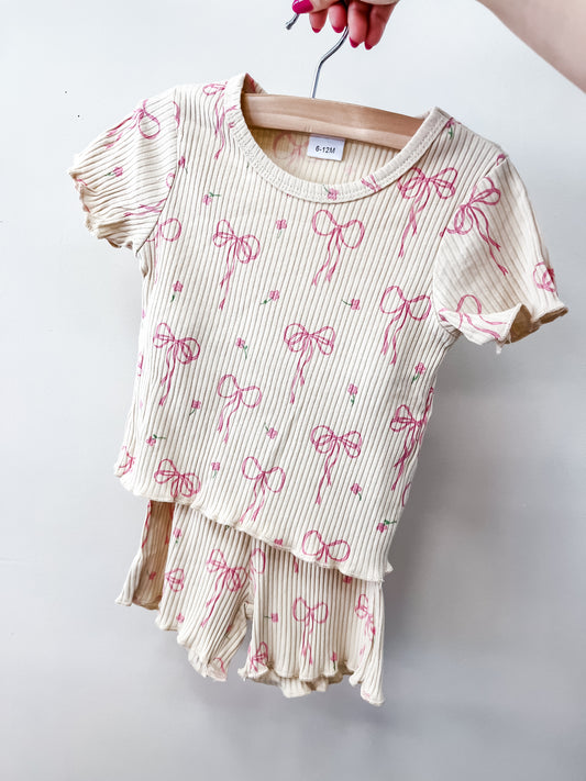 A hand displays the Sugar Bow Set by Tiny Tot Bundles—a cream-colored, ribbed two-piece kids' outfit with pink bows and green dots. The set includes a short-sleeve top and matching ruffled shorts.