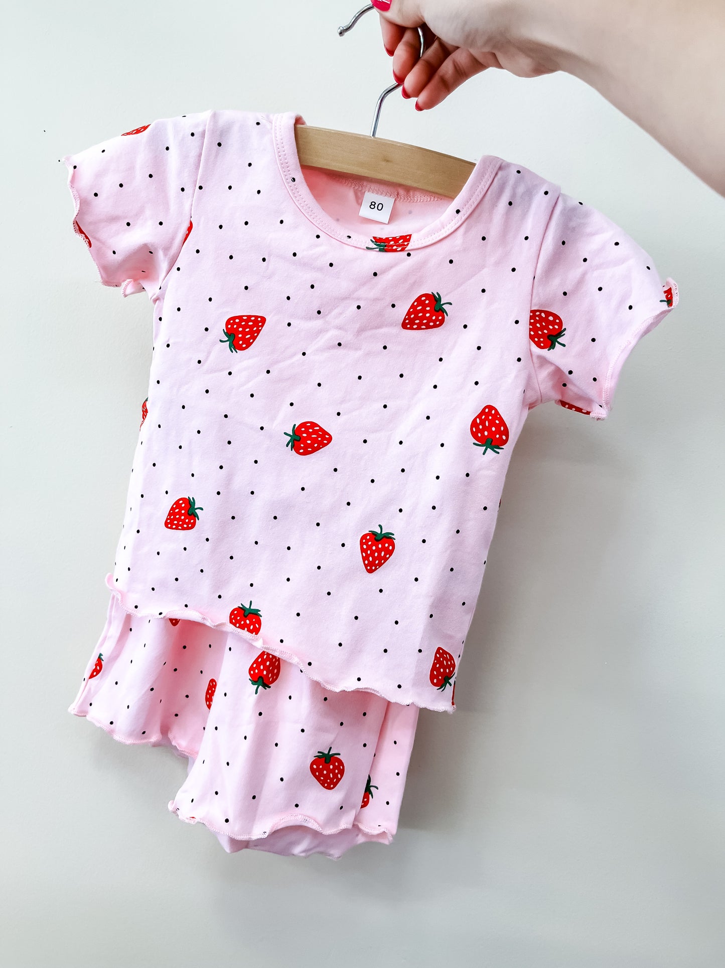 A hand displays the Berry Bliss Set by Tiny Tot Bundles—a baby pink short-sleeve shirt and shorts with black polka dots and red strawberry prints—on a wooden hanger against a plain white background.