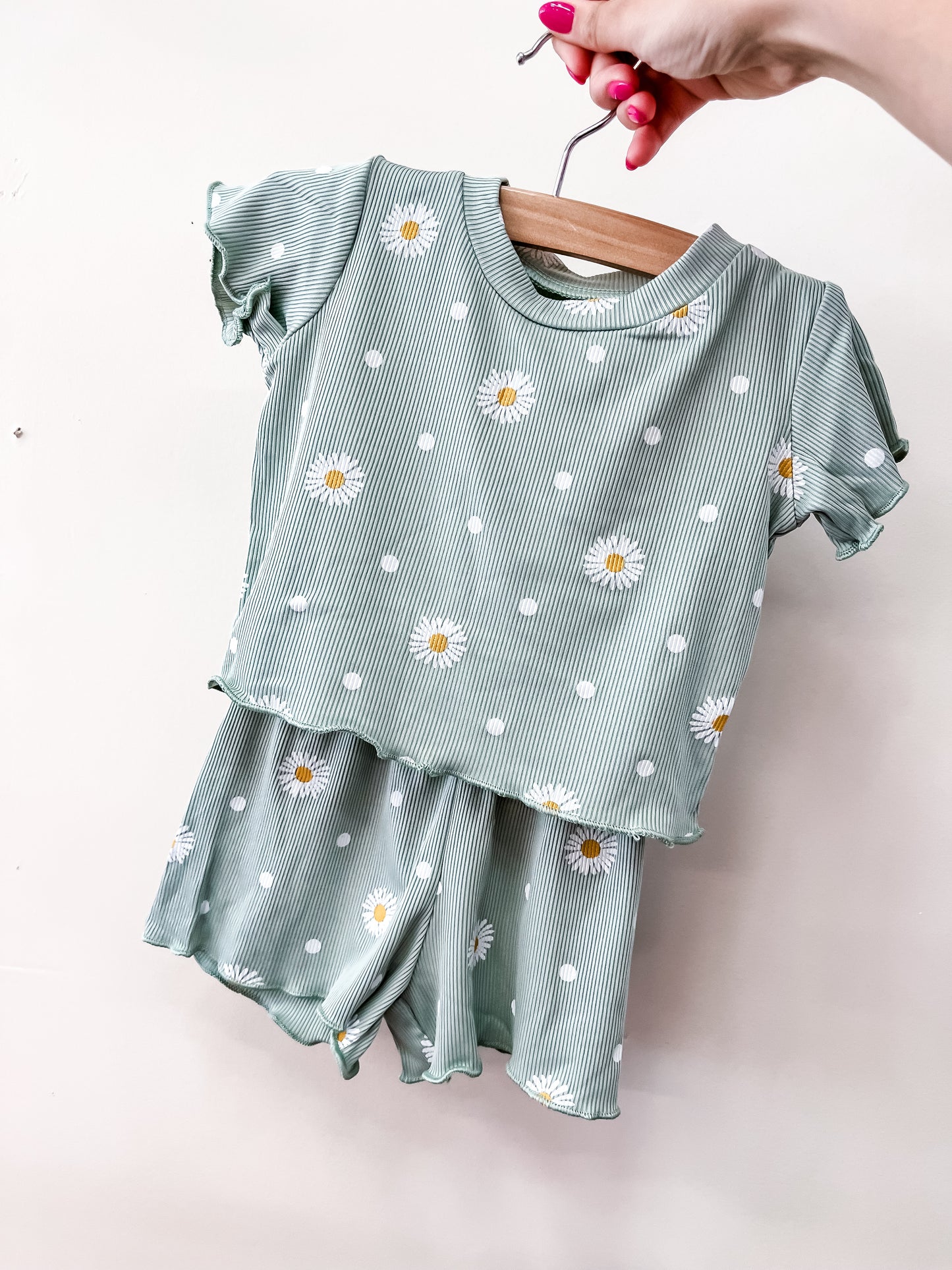 A hand holds up the Tiny Tot Bundles Minty Meadow Set—a green short-sleeve toddler outfit with white stripes and daisy prints—on a wooden hanger against a plain light background.