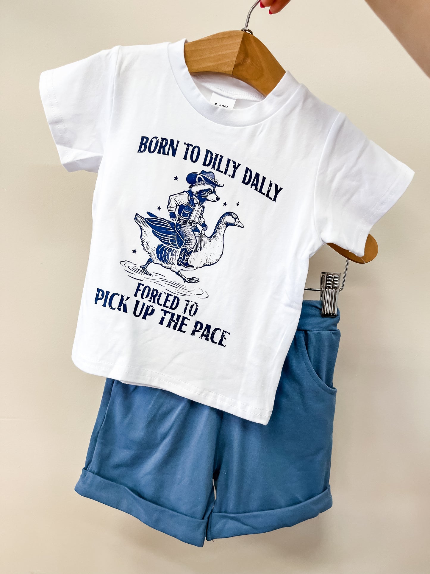 The Dilly Dally Set by Tiny Tot Bundles features a white children’s t-shirt with a duck riding a goose and the phrase "Born to Dilly Dally, Forced to Pick Up The Pace," paired with rolled blue shorts on a wooden hanger.