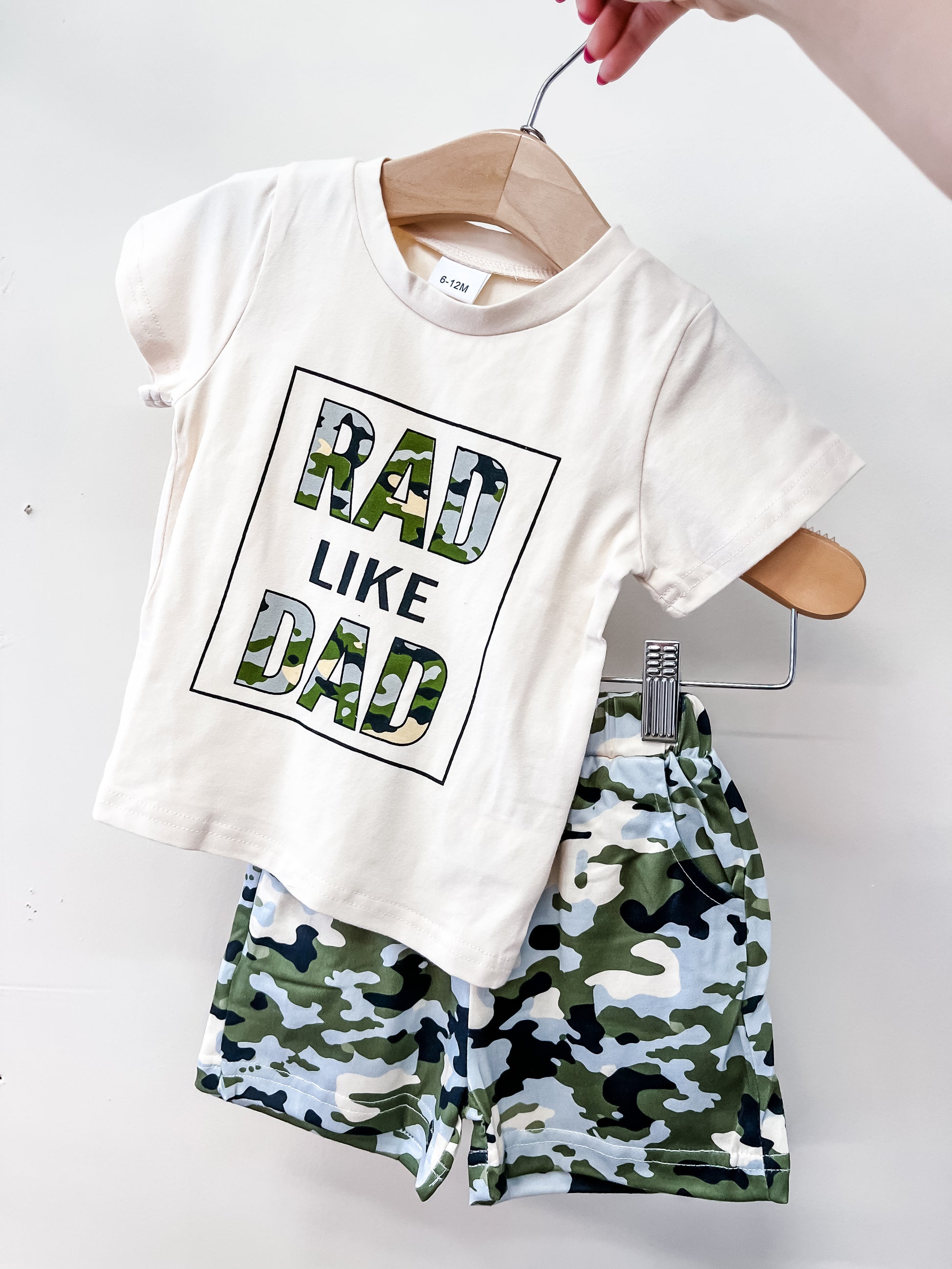 A hand holds Tiny Tot Bundles' Rad Like Dad Set: a cream t-shirt with "RAD LIKE DAD" in camo print, matched with green camouflage shorts.