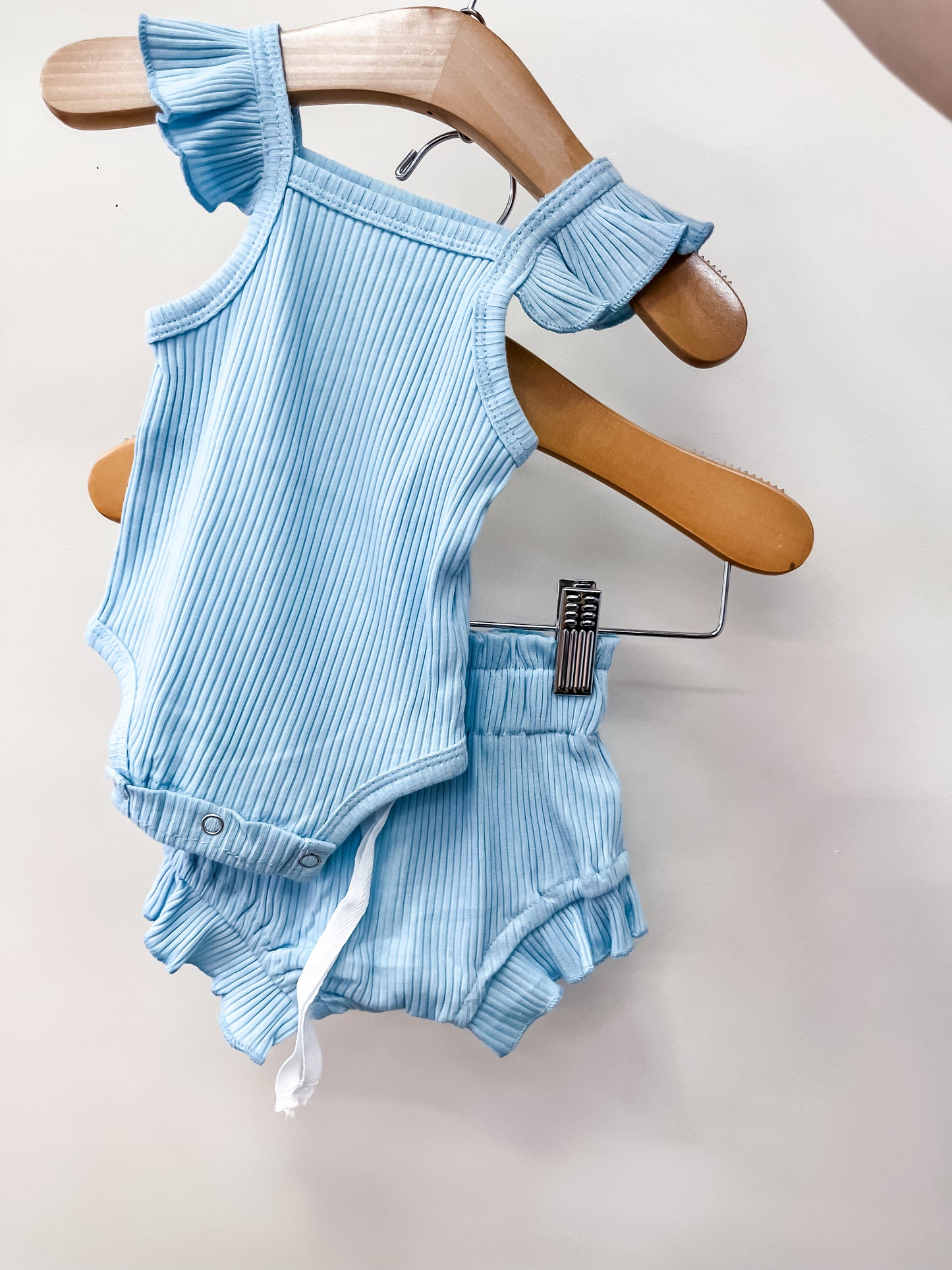 The Soft Sky Set by Tiny Tot Bundles features a light blue, ribbed sleeveless ruffled onesie with matching shorts and a white drawstring, both displayed on wooden hangers against a plain light background.