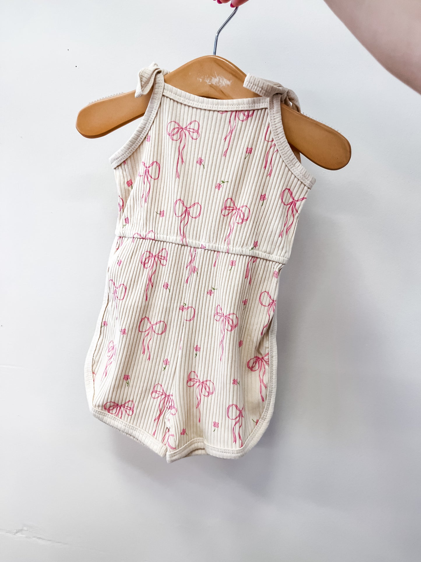 The Coquette Dreams Romper by Tiny Tot Bundles is a sleeveless baby romper with pink bows and stripes, shown on a wooden hanger against a white background.